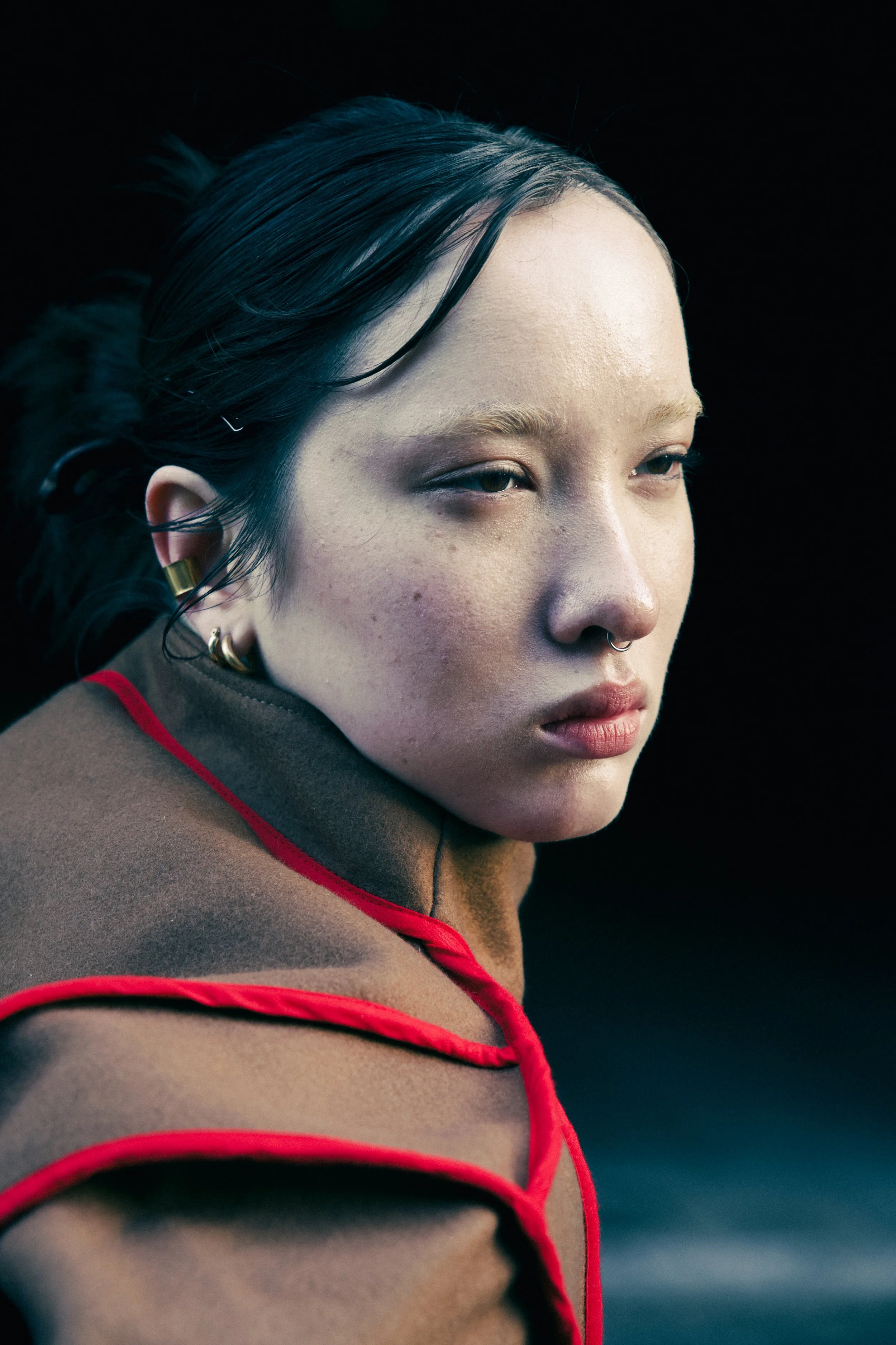 Close-up portrait of a young woman with short, dark hair, wearing gold earrings, and a septum piercing. She has light skin with freckles and is dressed in a brown top with red trim. The background is black.
