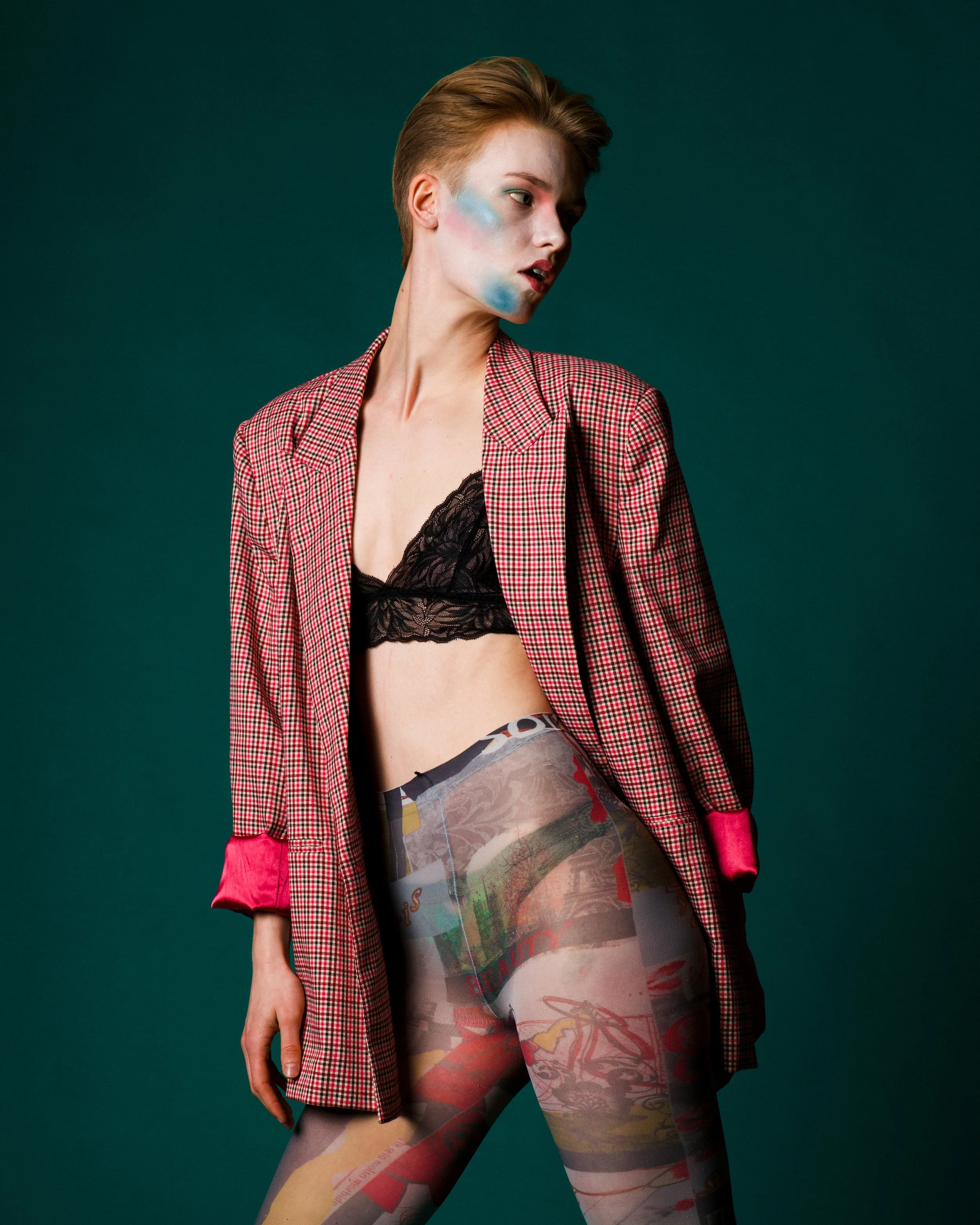 Fashion model with short red hair wearing a red checkered blazer, black lace bralette, and colorful printed tights, posing against a dark green background.