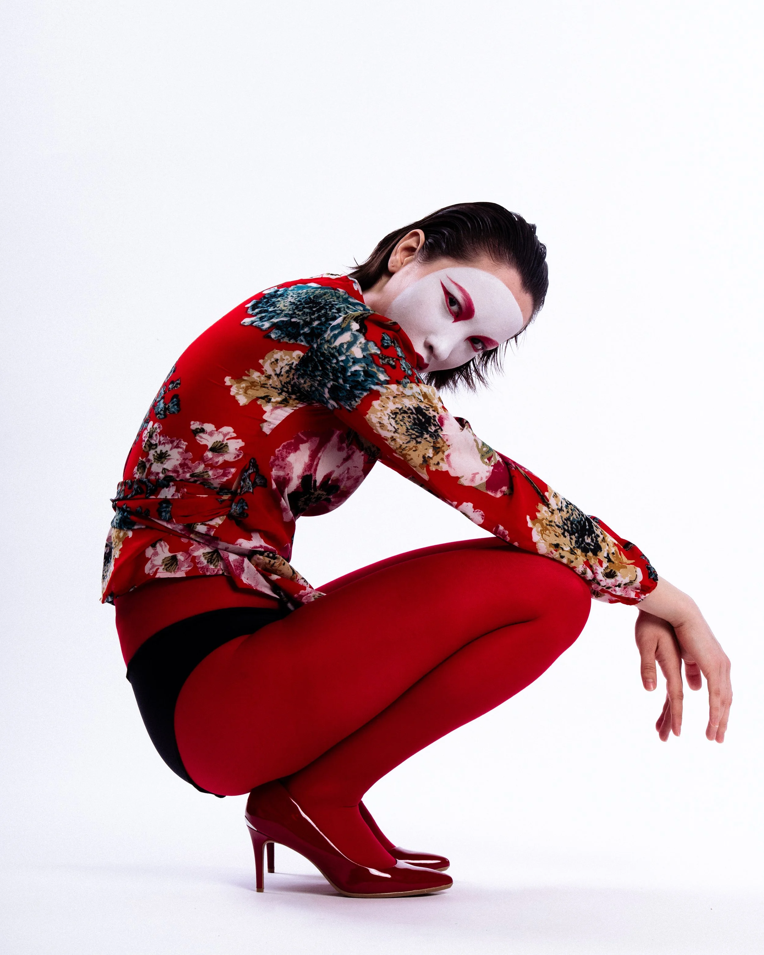 Person crouching in red tights, red high heels, and a floral top with a white face mask, posing against a white background.