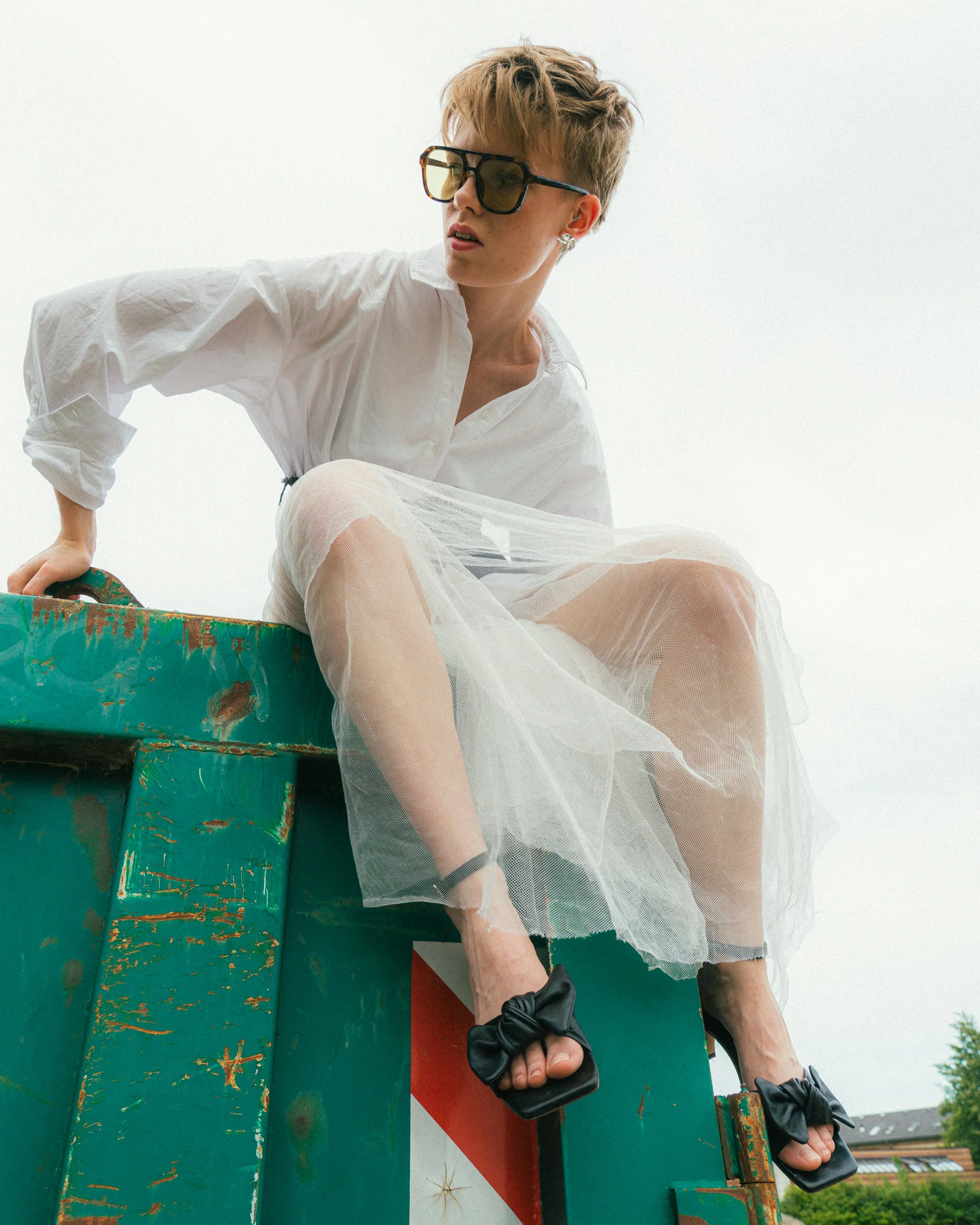 Woman sitting on a green, rusty metal structure outdoors, wearing oversized sunglasses, a white button-up shirt, a sheer tulle skirt, black high heels with bow details, and earrings.
