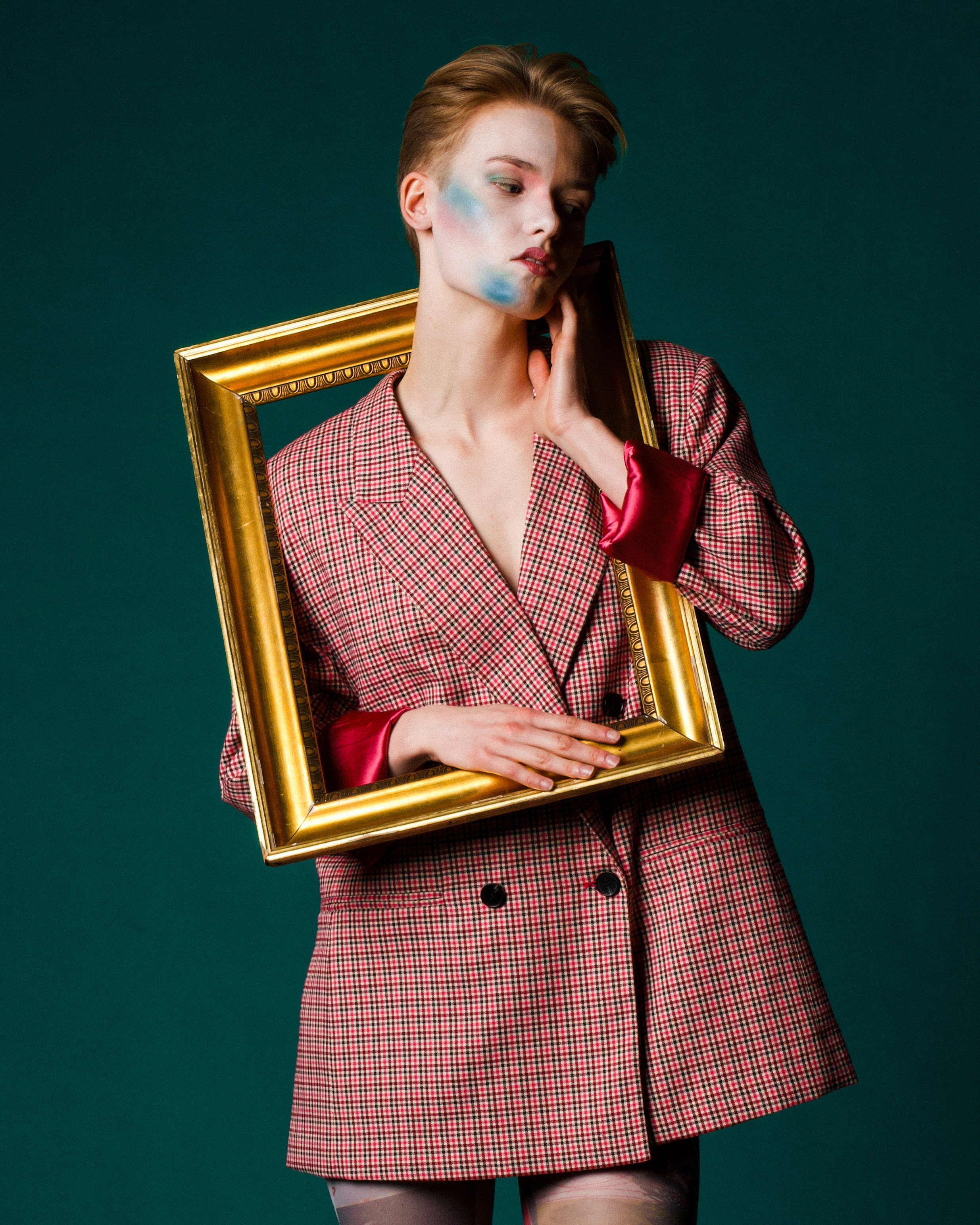 A woman with short hair wearing a red and black checkered blazer with red satin cuffs, holding a gold picture frame around her shoulders, standing against a teal background.