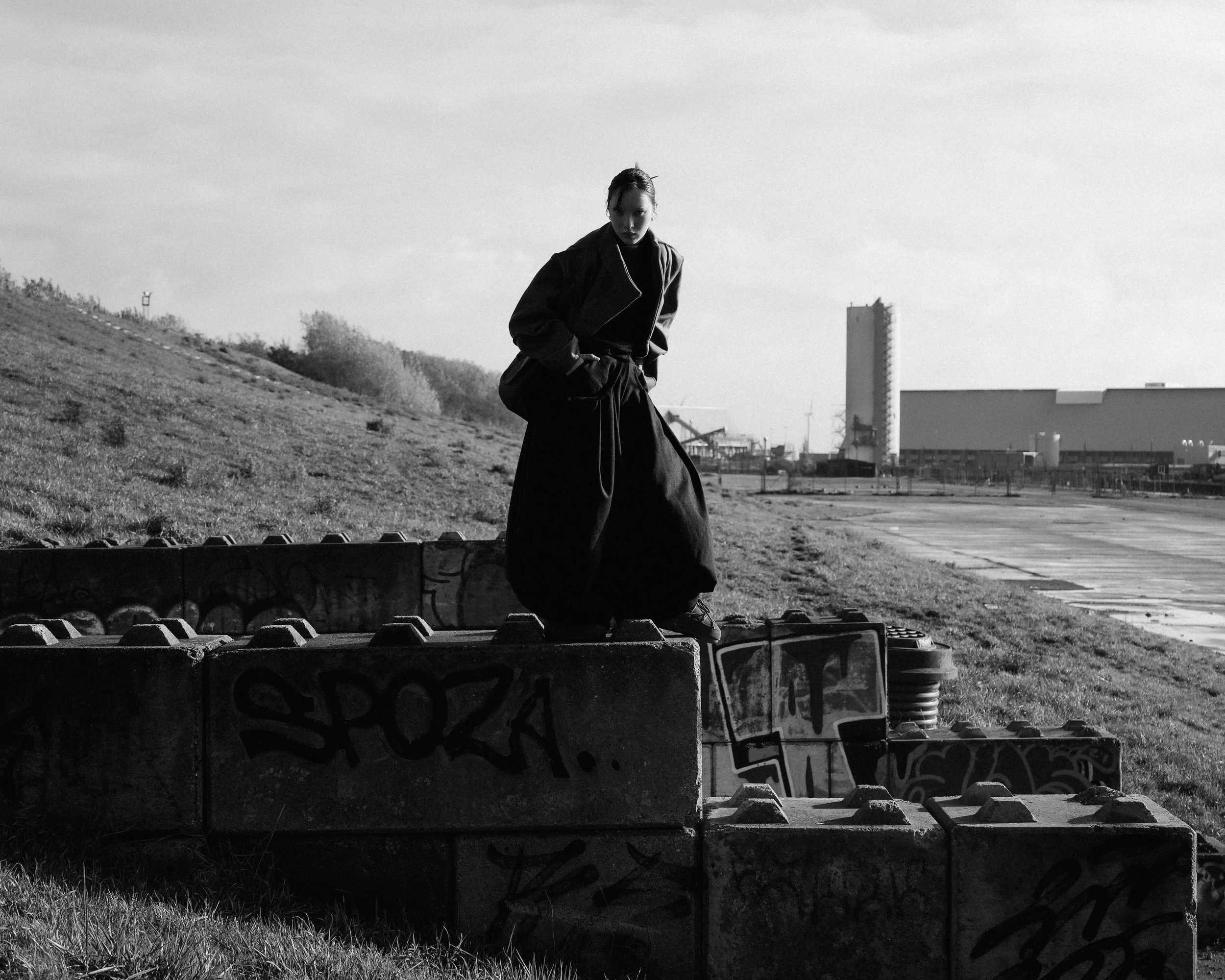 A person standing on graffiti-covered concrete blocks outdoors, with a grassy hill and industrial buildings in the background, black and white photo.