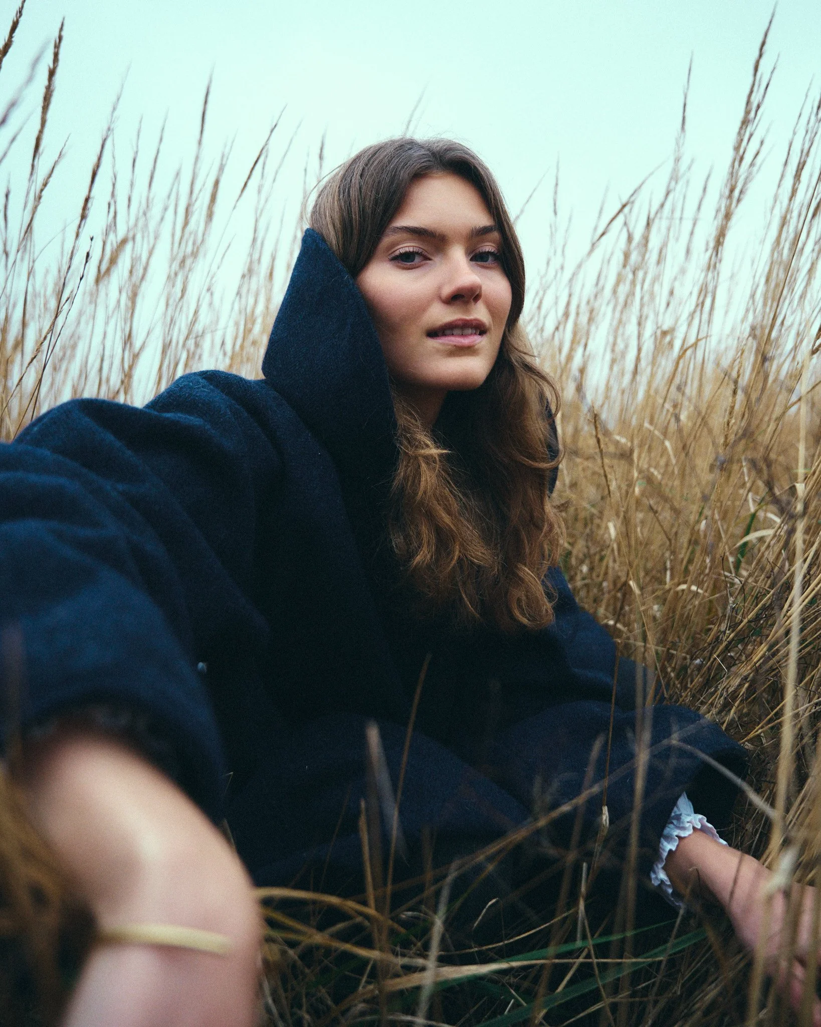 A young woman with brown hair wearing a blue coat sitting in tall beige grass during overcast weather.