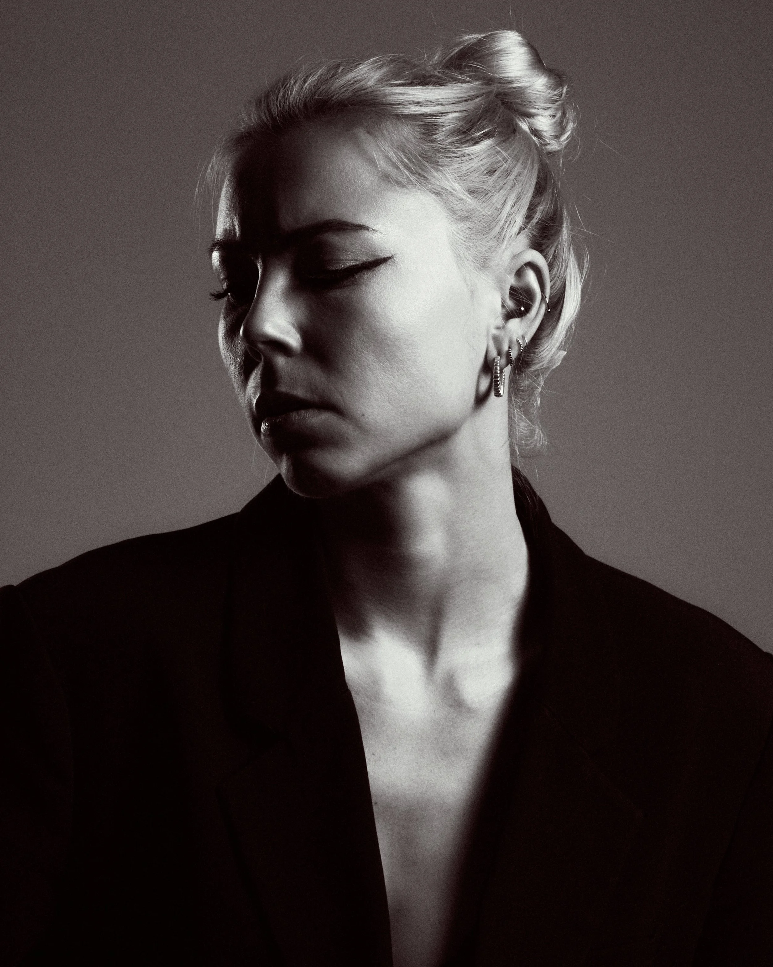 A black-and-white portrait of a woman with blonde hair styled in a bun, wearing multiple earrings and a dark blazer, looking downward with a serious expression.