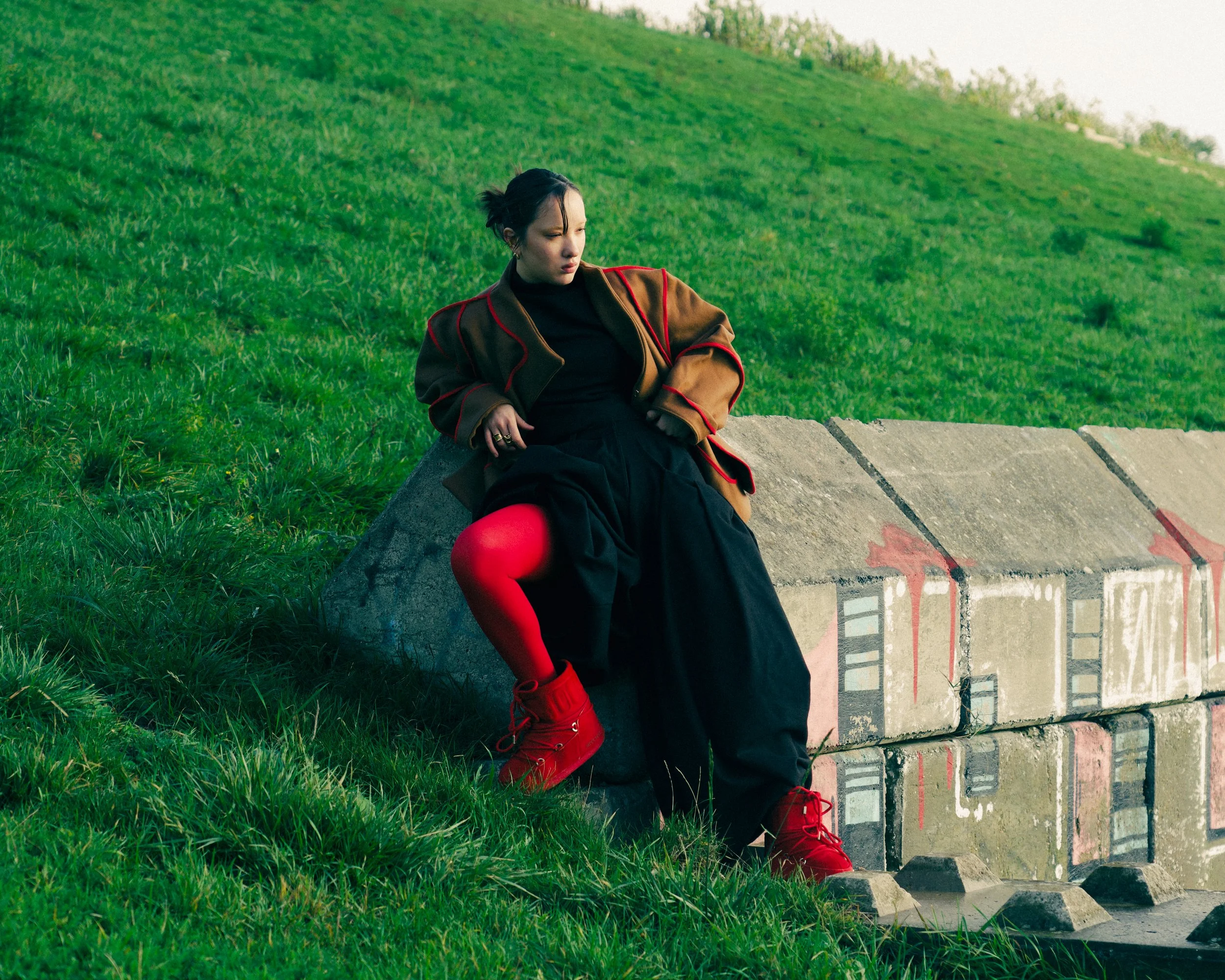 A woman sitting on a concrete block painted with cityscape graffiti in a grassy field during daytime, wearing a brown jacket, black dress, red tights, and red boots.