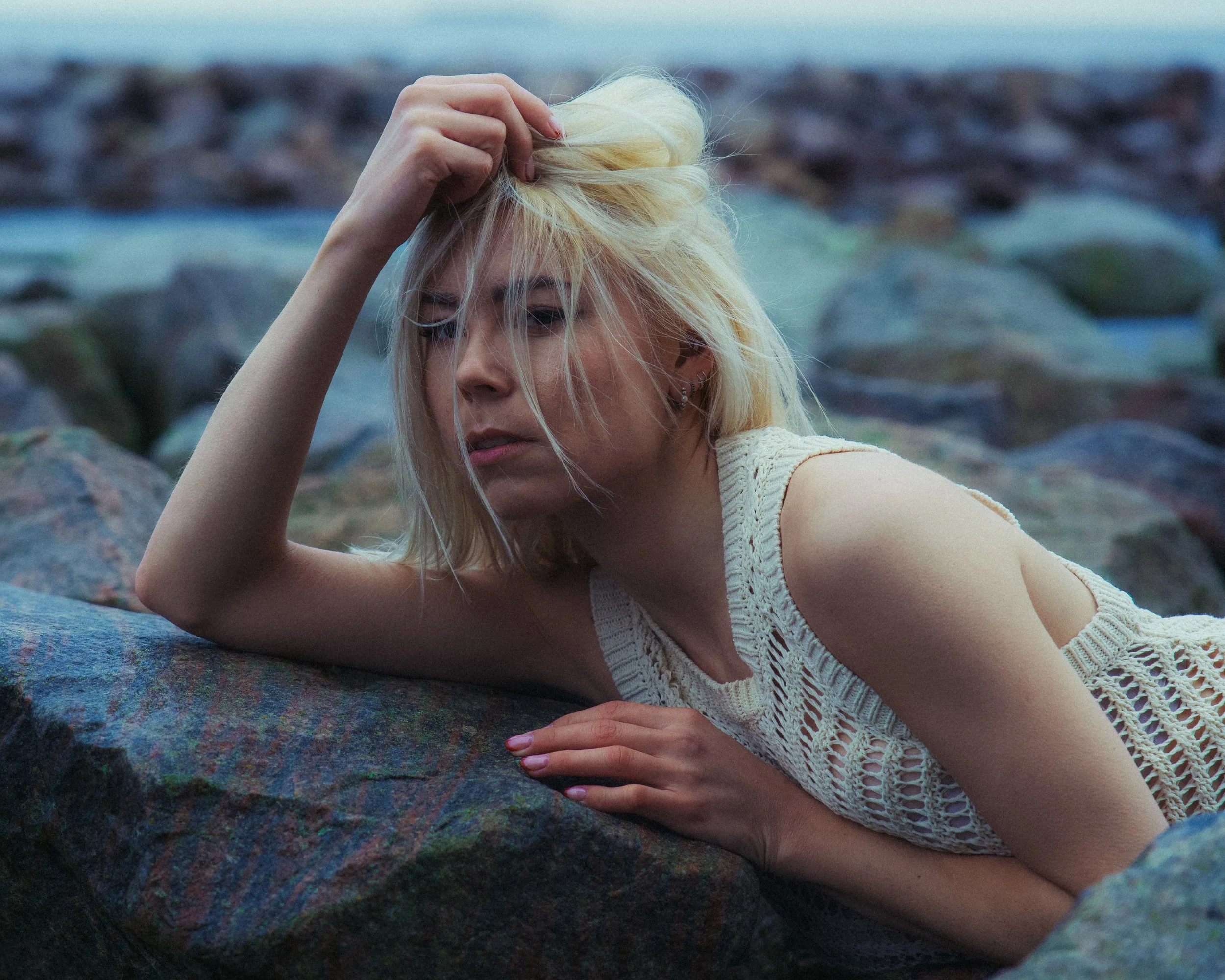 A blonde woman in a cream crochet dress is lying on rocks near water, with her eyes closed and hand running through her hair, appearing contemplative.