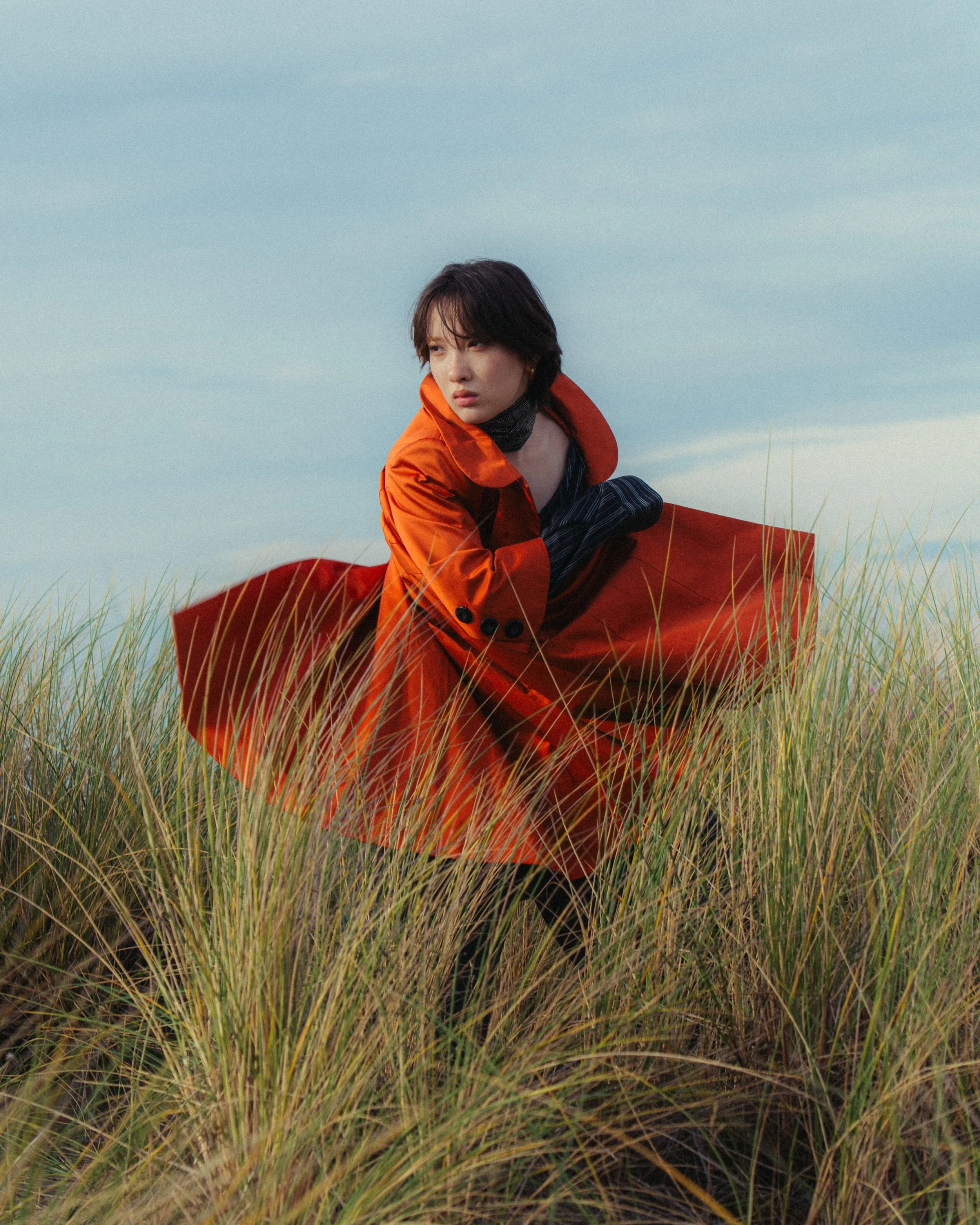 Woman in an orange coat crouching in tall grass with a cloudy sky background.