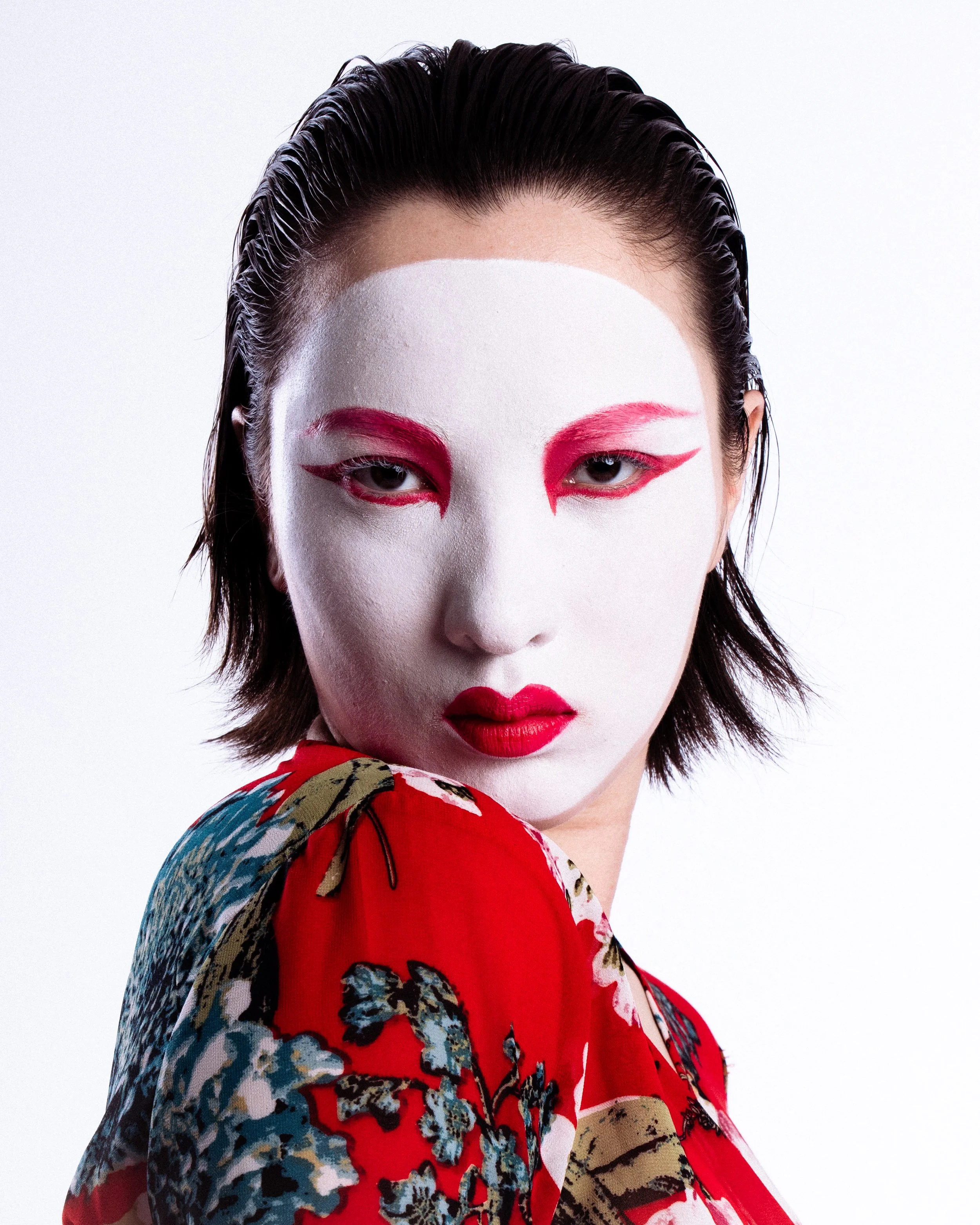 Close-up of a woman with traditional Japanese face makeup, including a white base, red and black eye makeup, and red lipstick, wearing a red floral kimono.