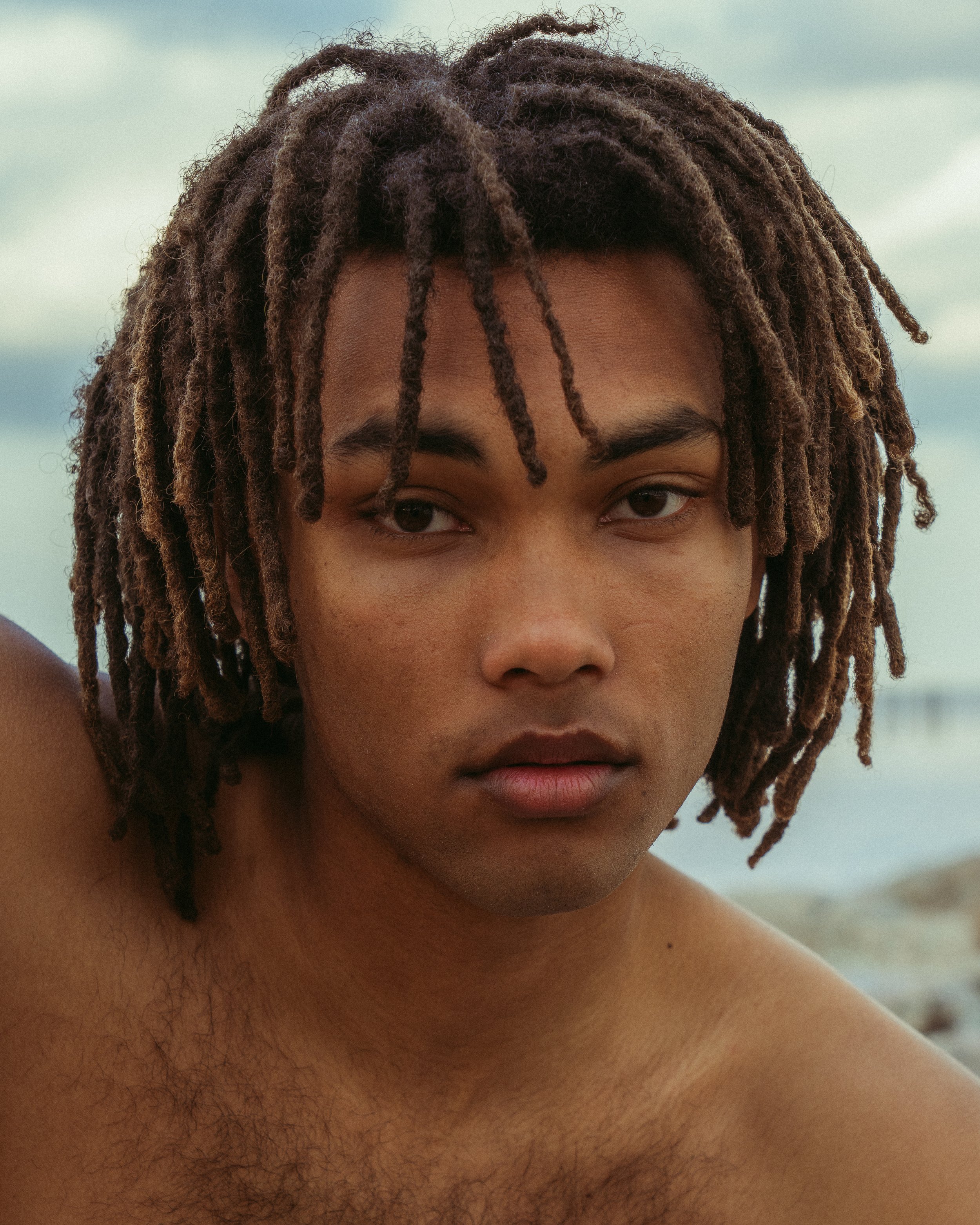 Close-up of a young man with dreadlocks at the beach, looking into the camera with a serious expression.