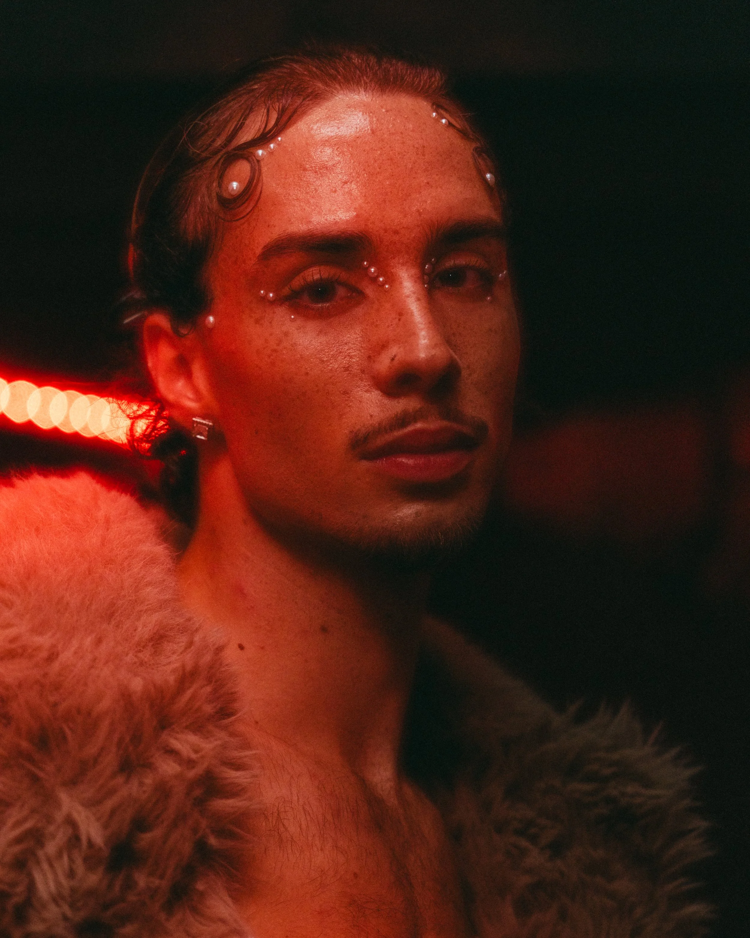 A person with a serious expression, wearing pearl and rhinestone embellishments on their face, with short hair, earrings, and a fur coat, in a dark setting with red lighting.
