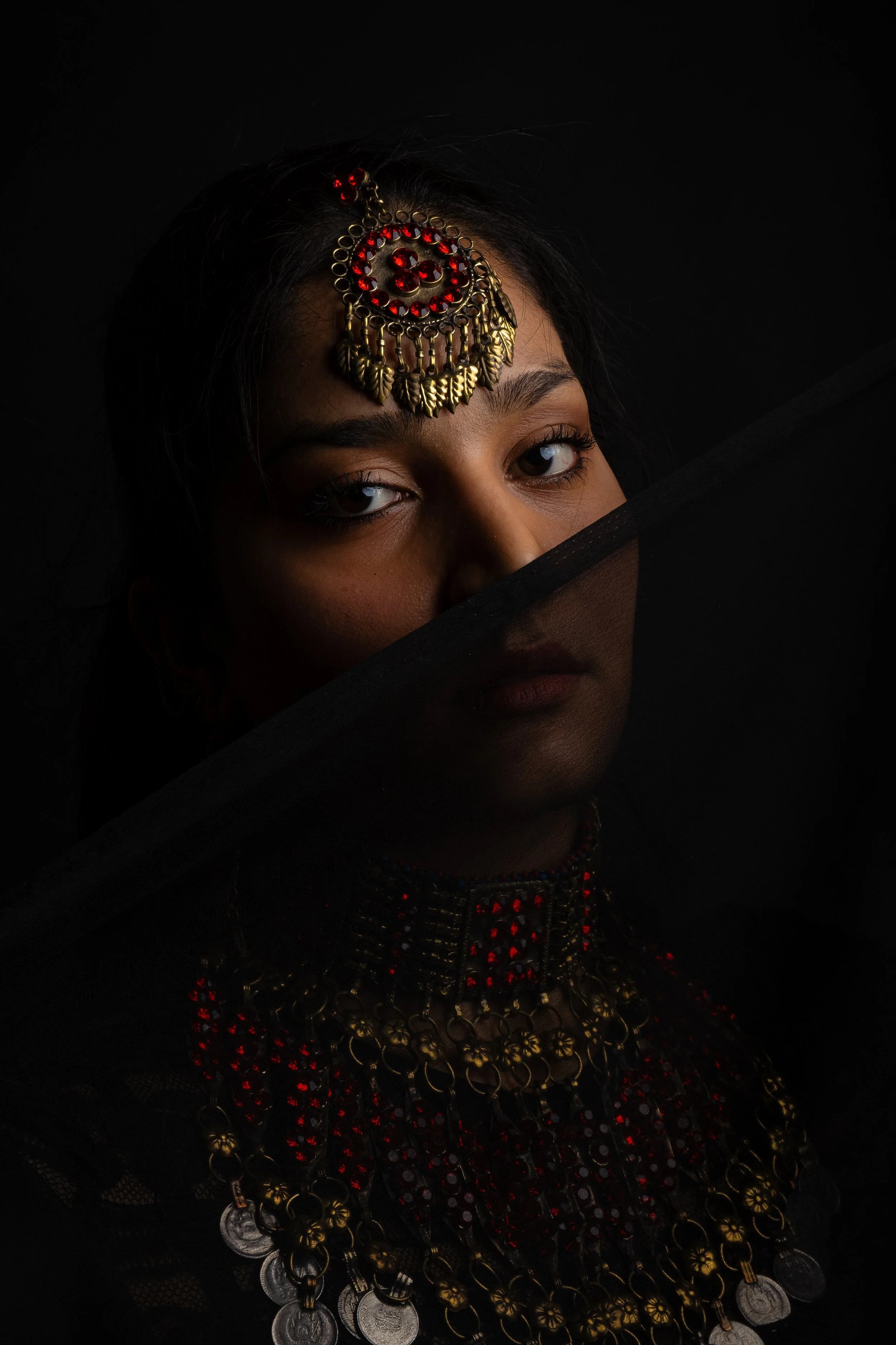 A woman wearing traditional jewelry, including a decorative headpiece and layered necklaces, partially obscured by a black sheer fabric, with a dark background.