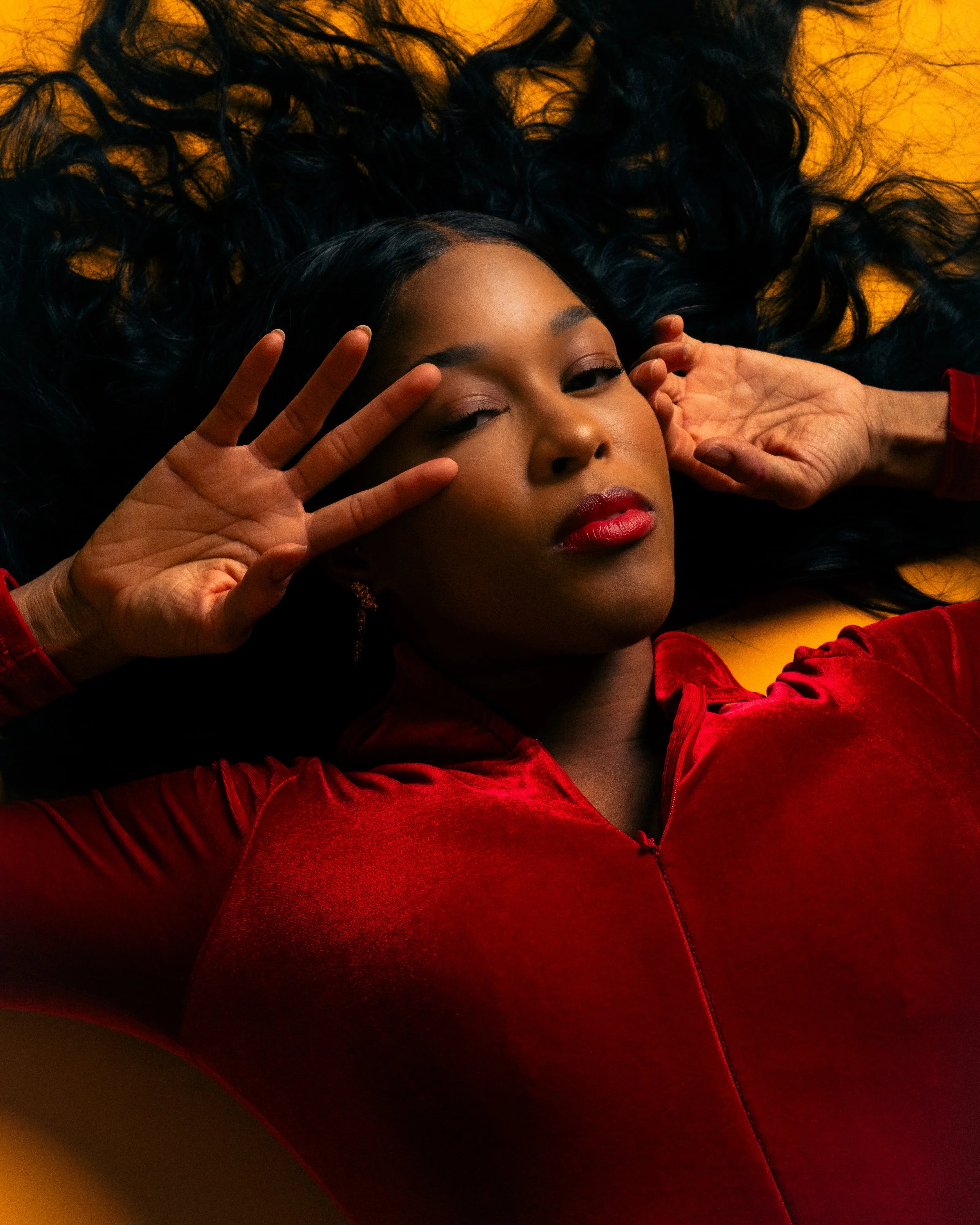 A woman with long black curly hair lying on a yellow background, wearing a red velvet top, with one eye closed and lips slightly parted.