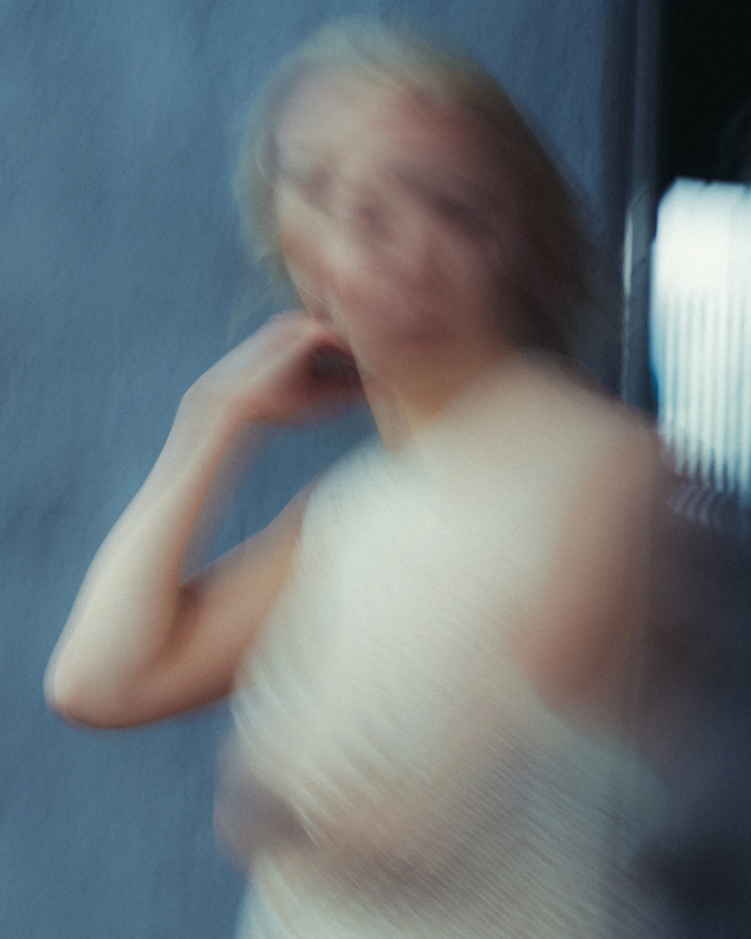 Blurry photo of a woman with blonde hair, wearing a white top, with her hand near her face, against a blue background