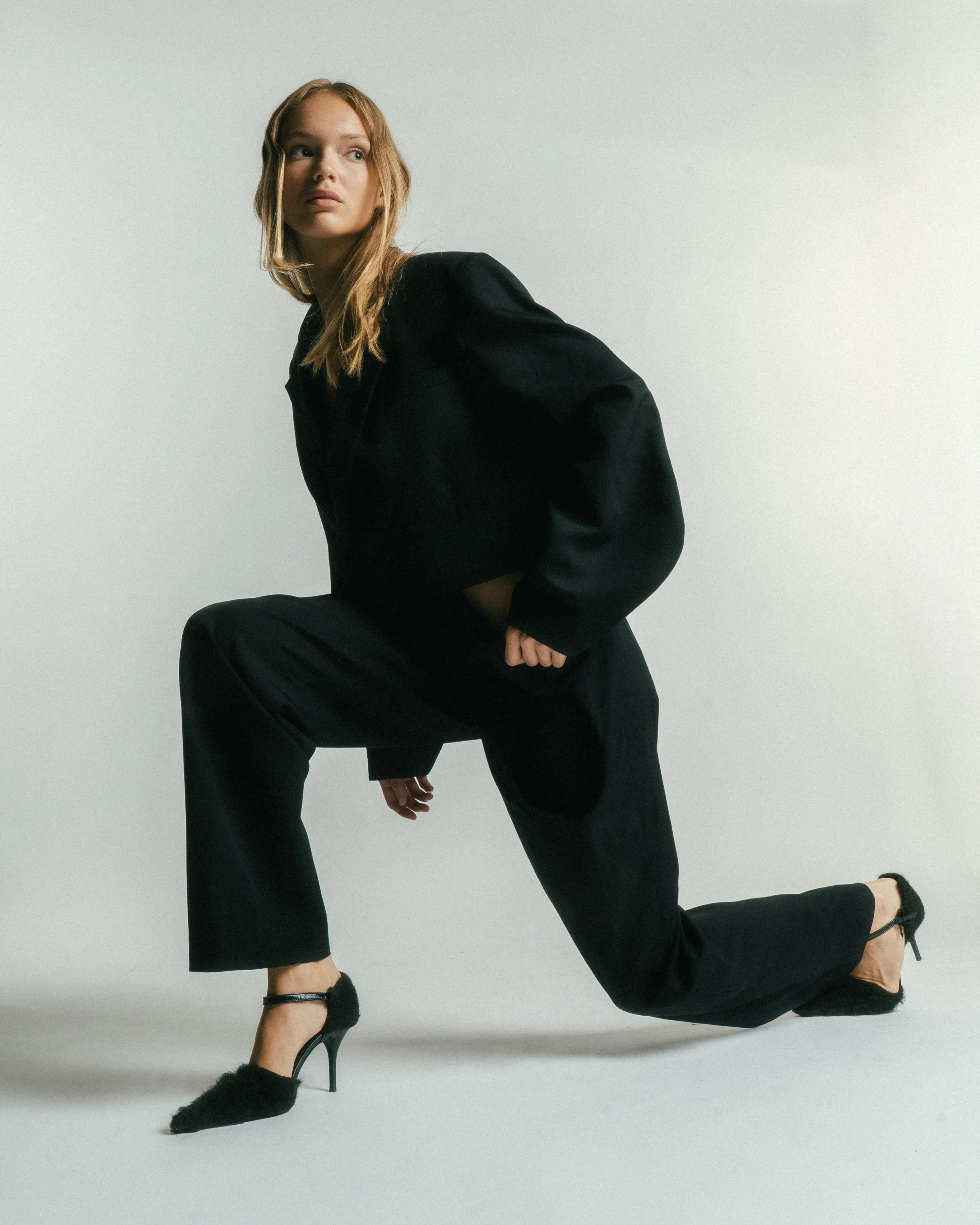 A woman with red hair in a black suit and high heels kneeling on one knee against a light background.