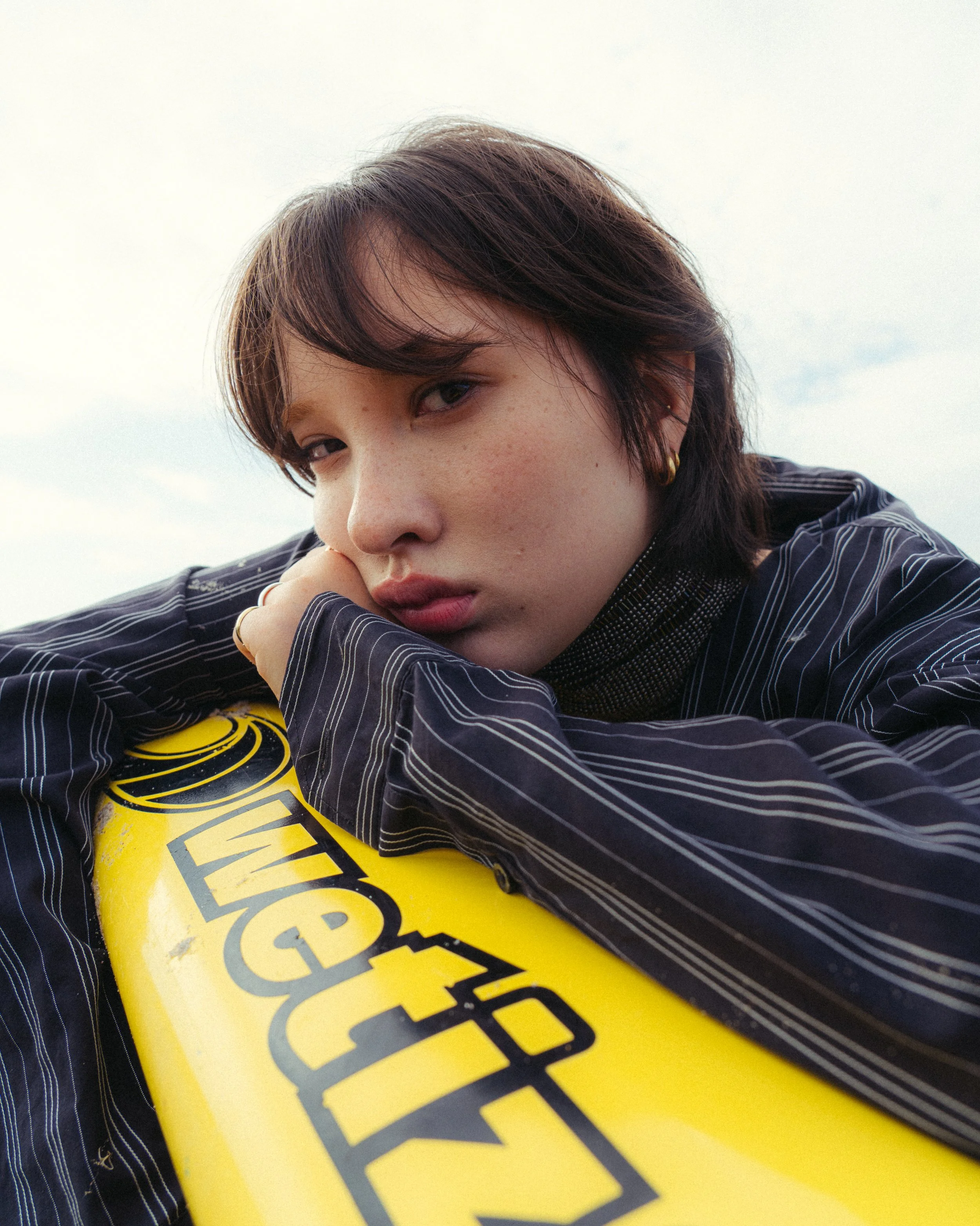 Close-up of a young woman resting her chin on her arm, leaning on a yellow surfboard with black lettering, outdoors on a cloudy day.