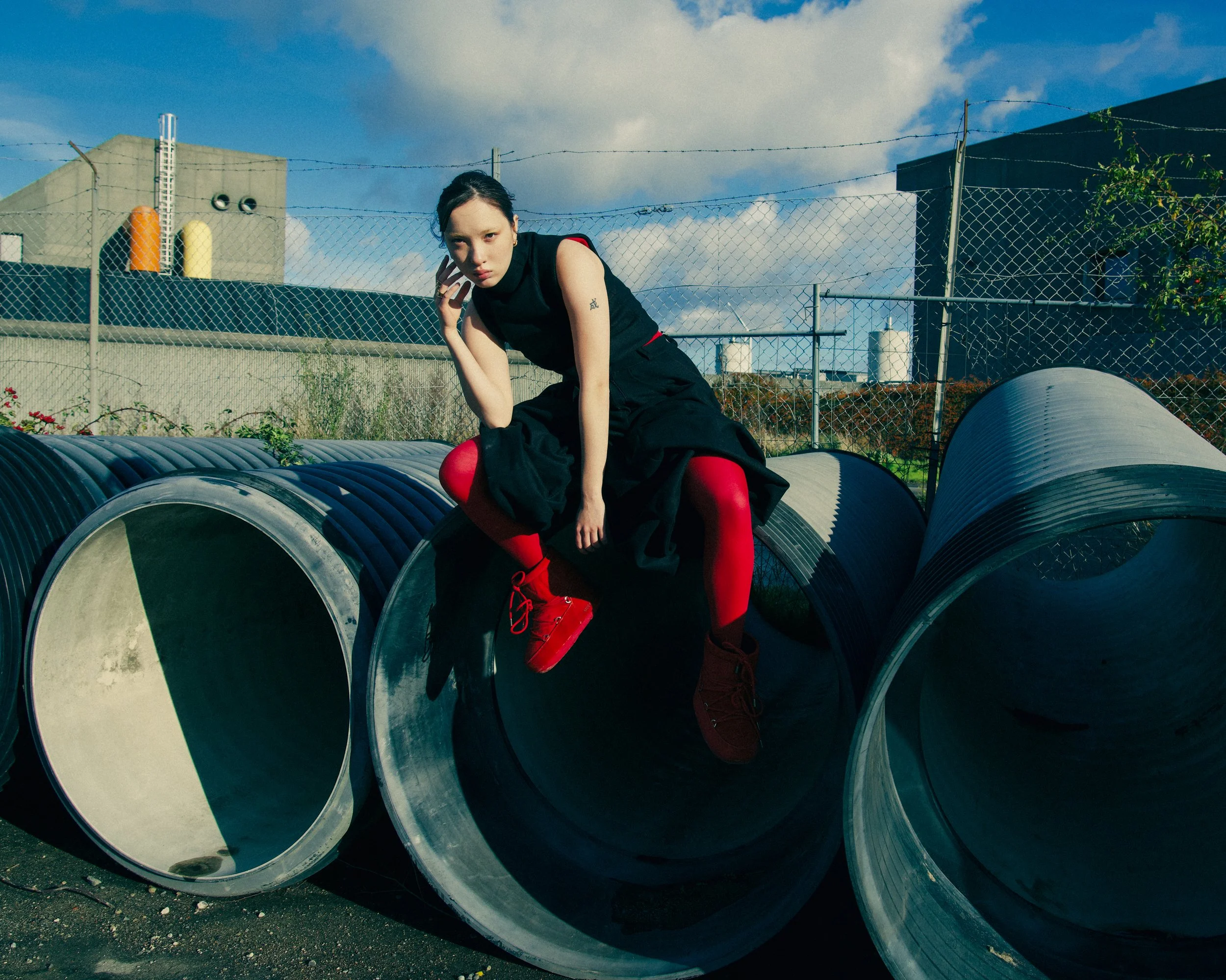 A woman with dark hair and fair skin sitting on large industrial pipes outdoors against a fence and industrial buildings under a blue sky with clouds. She is wearing a black sleeveless top, a black skirt, red tights, and red shoes.