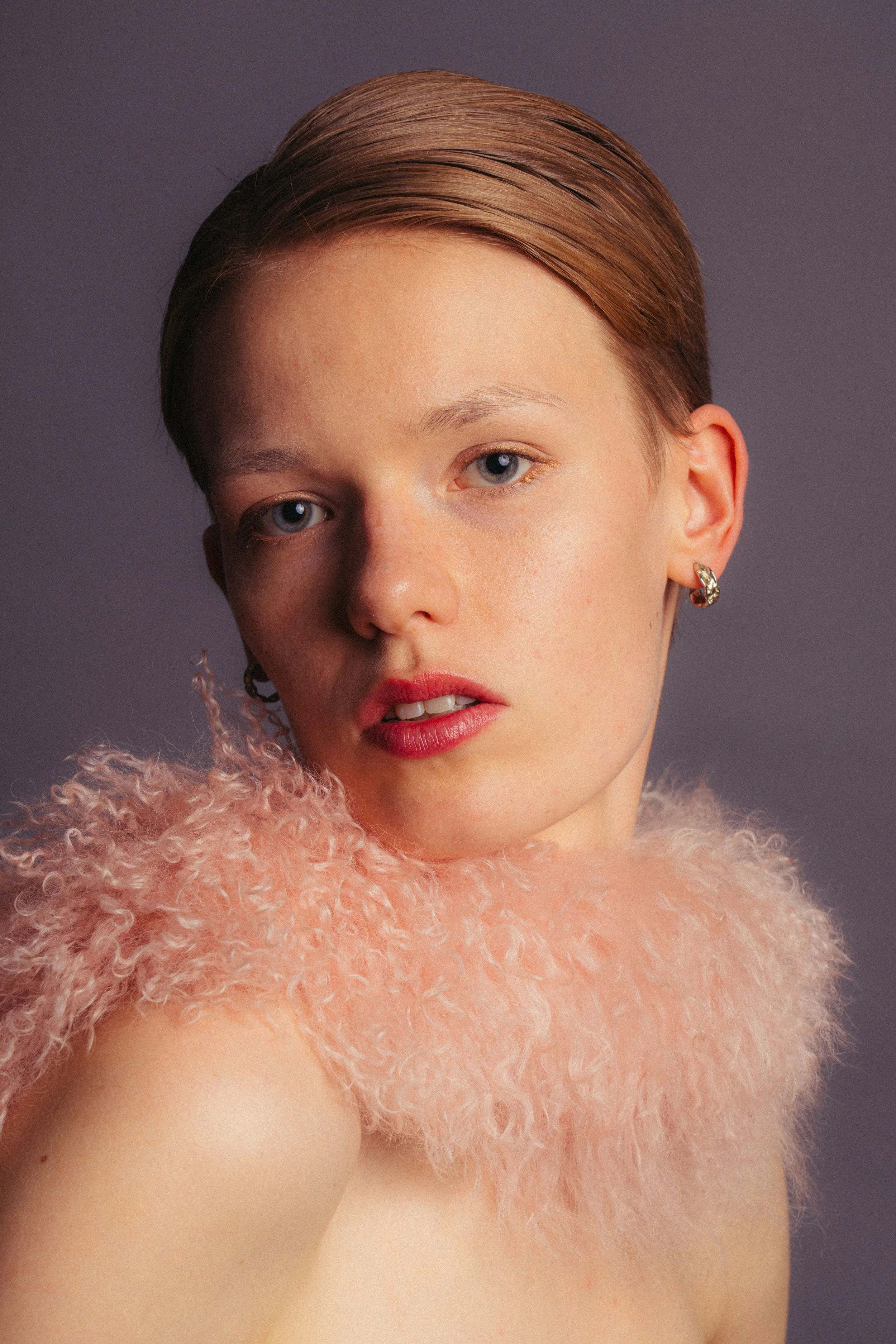 A young woman with short, slicked-back hair and blue eyes wearing pink lipstick, earrings, and a fluffy pink garment.