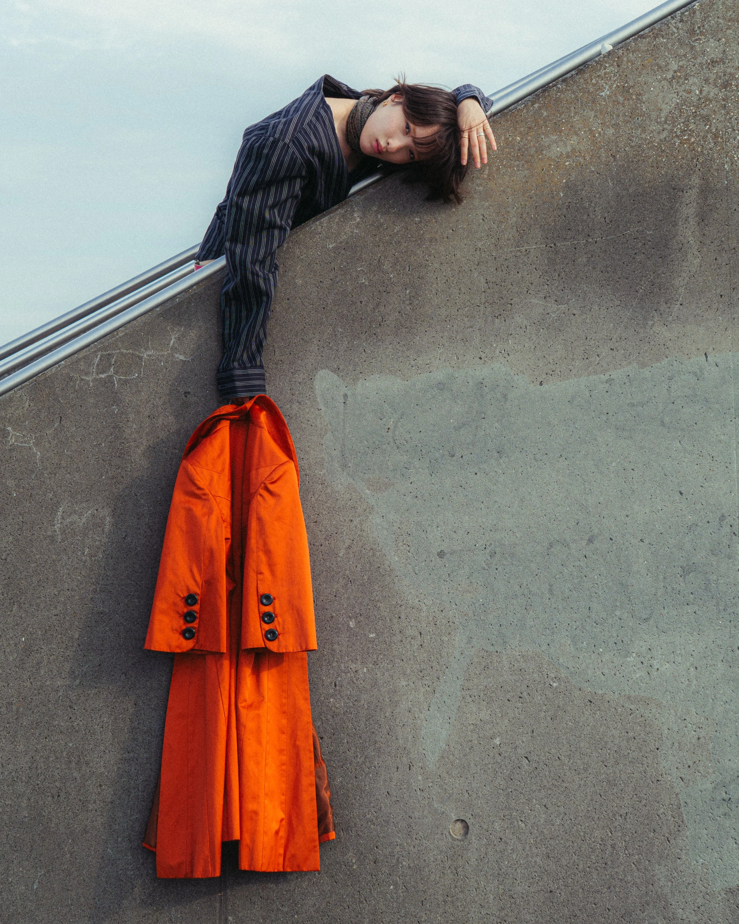 A woman lying on her side on a concrete surface, supporting her head with her hand, with an orange jacket hanging on the wall below her.