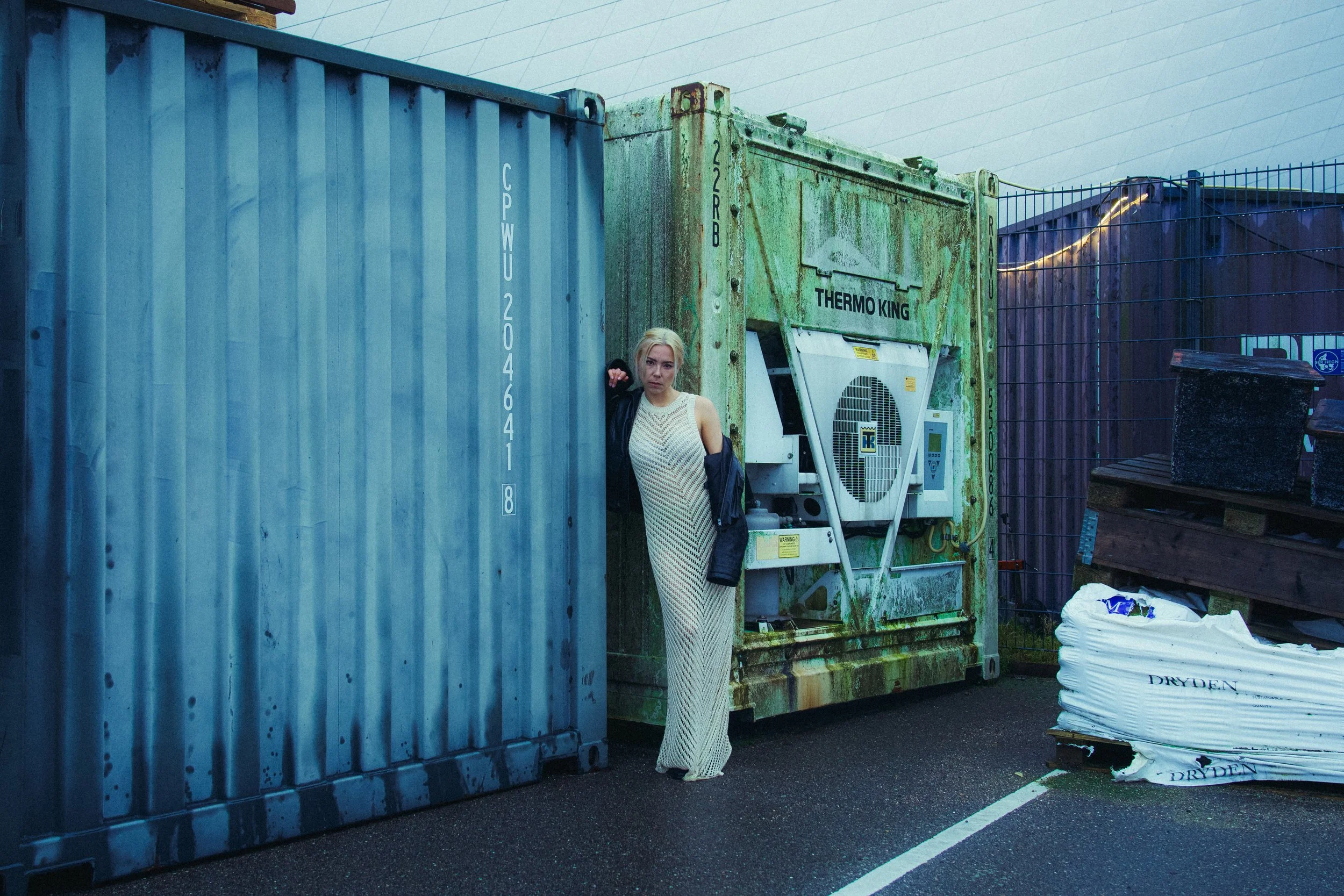 A woman in a long, sleeveless dress standing between two large dumpsters in an outdoor setting. The woman has blonde hair and is wearing a black jacket draped over her shoulders.