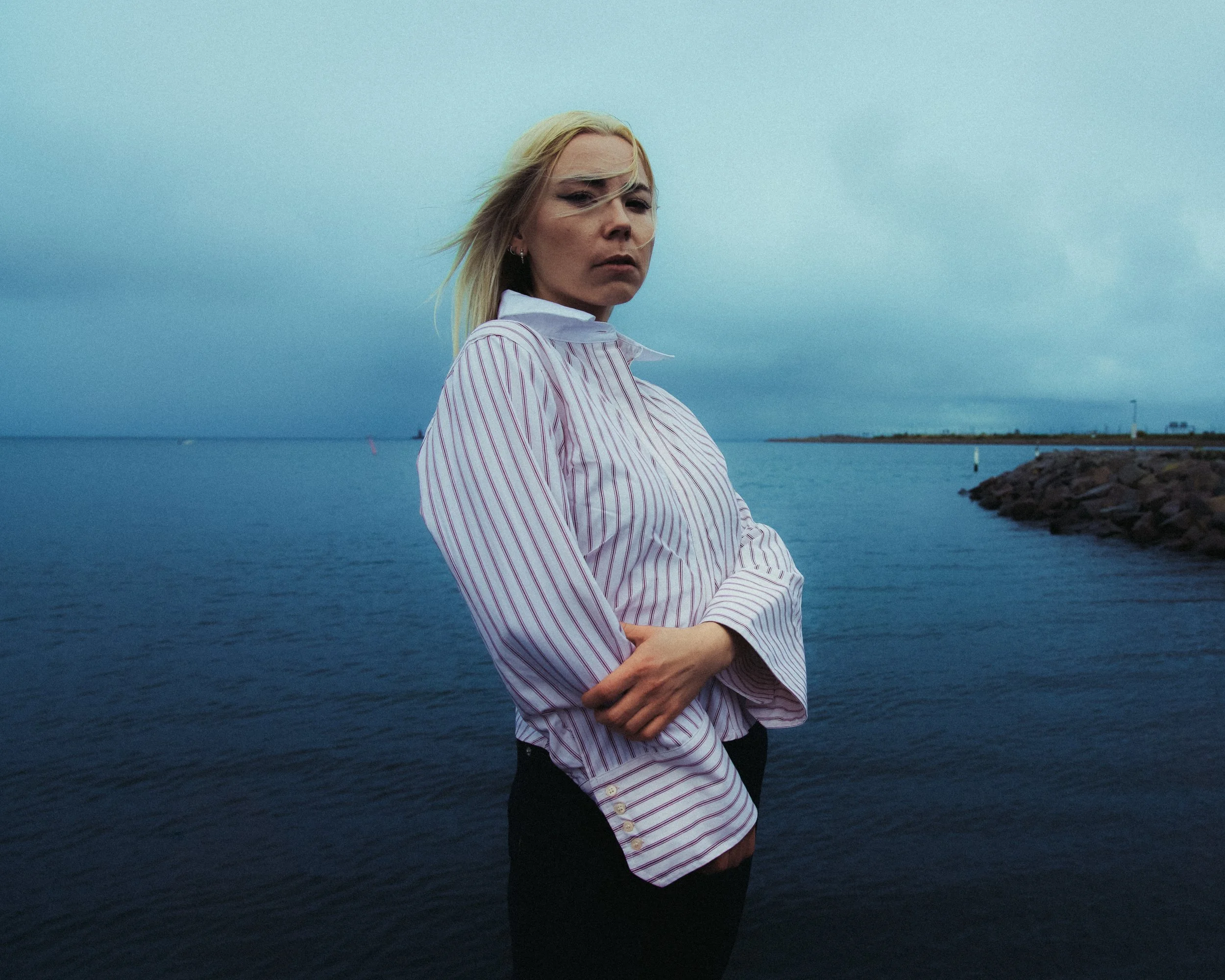 A young woman with blonde hair standing near water under a cloudy sky, wearing a white and red striped shirt with her arms crossed.