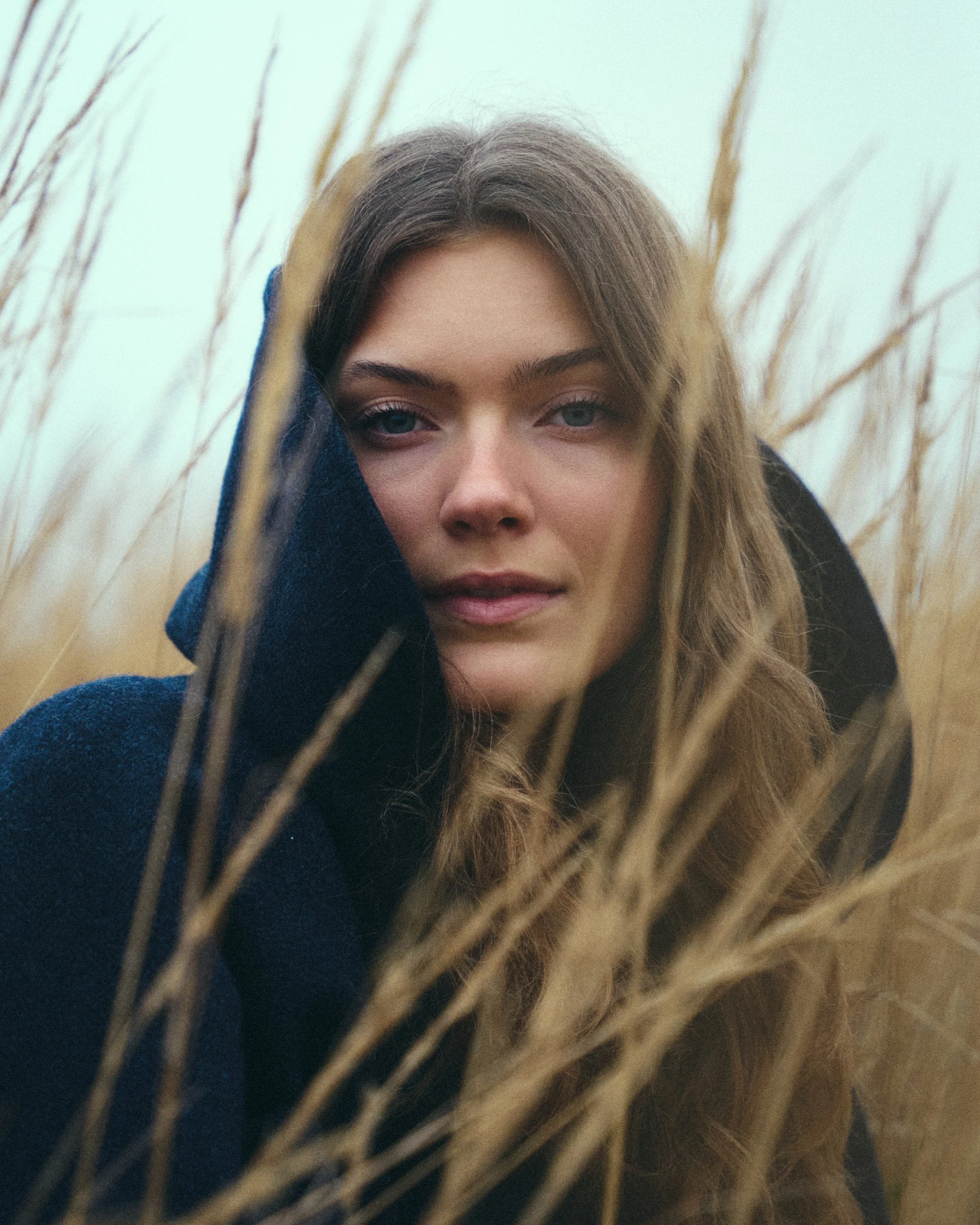 A young woman with long wavy hair and blue eyes in a field of tall, dried grass, wearing a dark hoodie, looking directly at the camera.