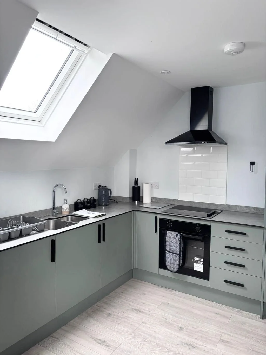 Modern kitchen with light green cabinets, a black oven with a towel, gray countertop, and a black range hood. There is a skylight window in the ceiling and small kitchen appliances on the counter.