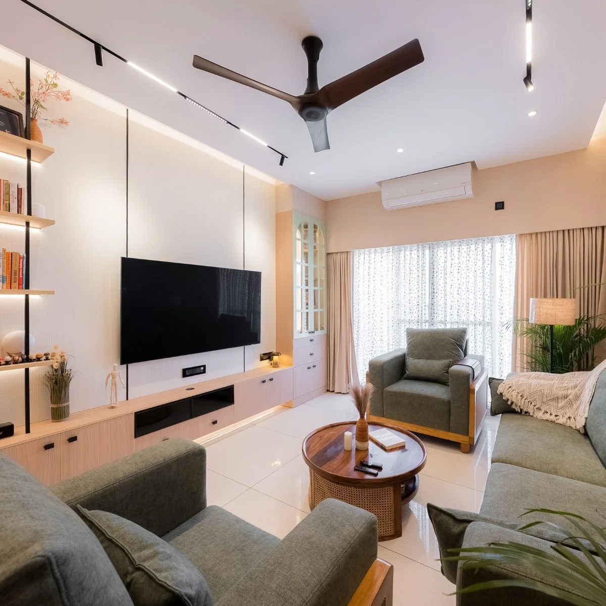 Modern living room with a ceiling fan, wall-mounted TV, beige curtains, green sofas, round wooden coffee table, bookshelf, and plants.