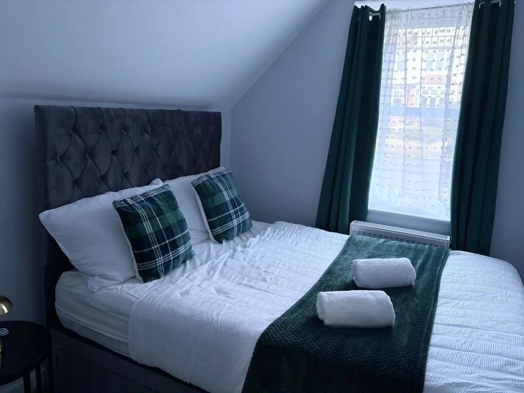A neatly made bed with white sheets, green plaid pillows, and two rolled white towels on a dark green blanket in a bedroom with green curtains and a window with a city view.