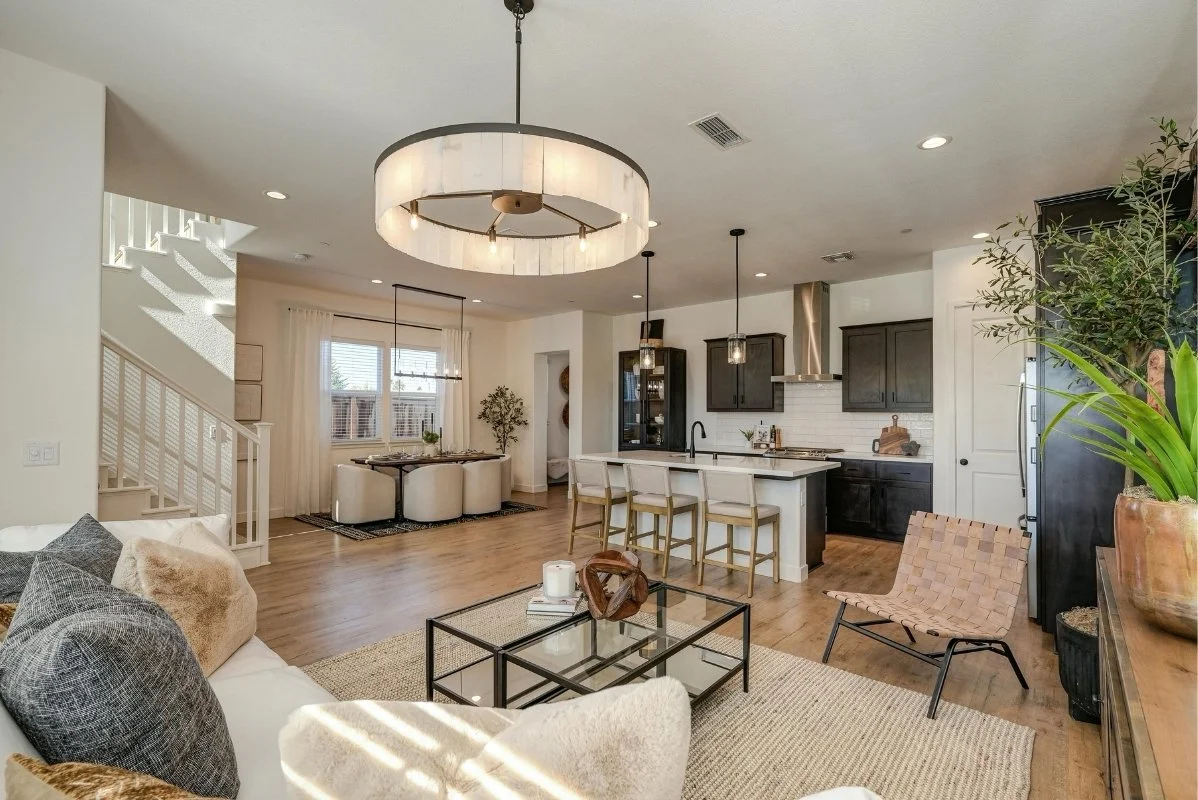 Open-concept living room and kitchen with modern decor, a large ceiling light, bar stools at the kitchen island, a dining table near a window, and various plants.