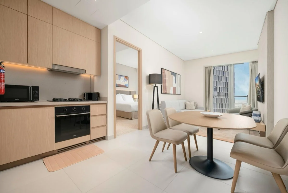 Modern hotel studio featuring a kitchenette with wooden cabinets and black appliances, adjacent to a dining area with a round table and four beige chairs, with a view of a bedroom and large window with city view in the background.