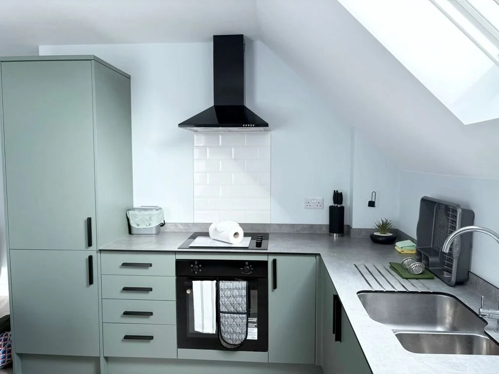 Modern kitchen with gray cabinets, a black range hood, white subway tile backsplash, stovetop, oven, sink, and minimal decor under a sloped ceiling with a skylight.
