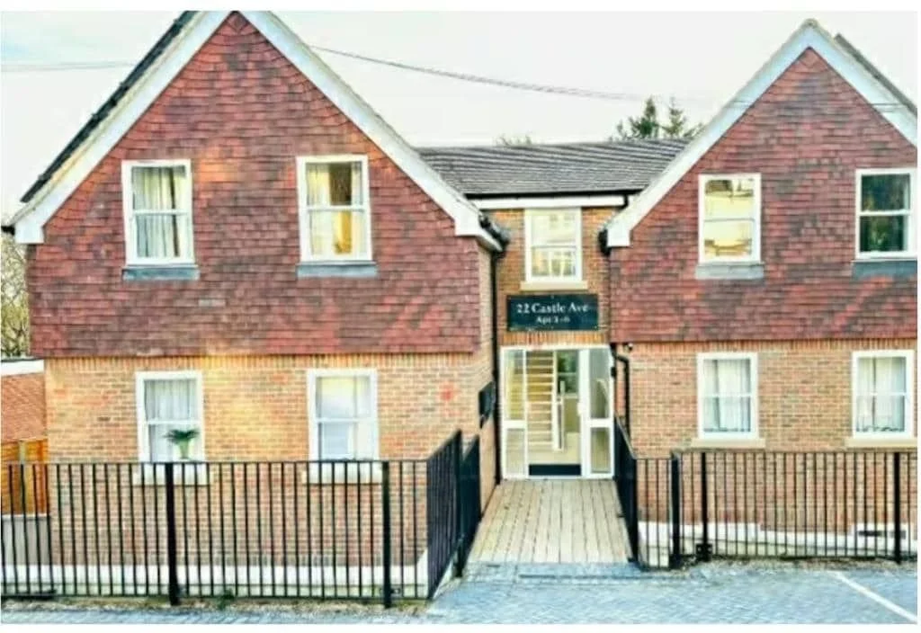 A two-story brick building with a red shingled gable roof, featuring four windows with white curtains, a black sign reading '22 Castle Ave Apt 4-8', and a wheelchair-accessible ramp leading to the entrance, enclosed by black metal fencing.