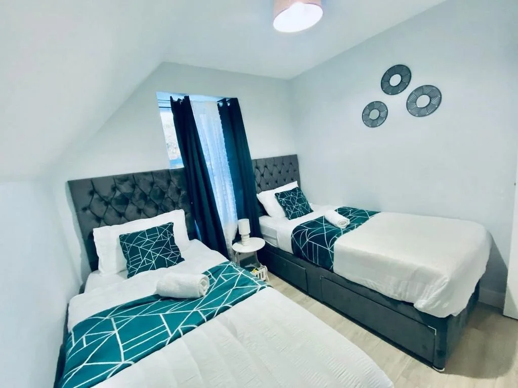 Two beds with tufted grey headboards, white bedding, and blue geometric decorative pillows and runners. A small white nightstand with a lamp between the beds. A window with dark blue curtains and a window covering, three decorative circular wall hangings on the white wall, and a ceiling light.