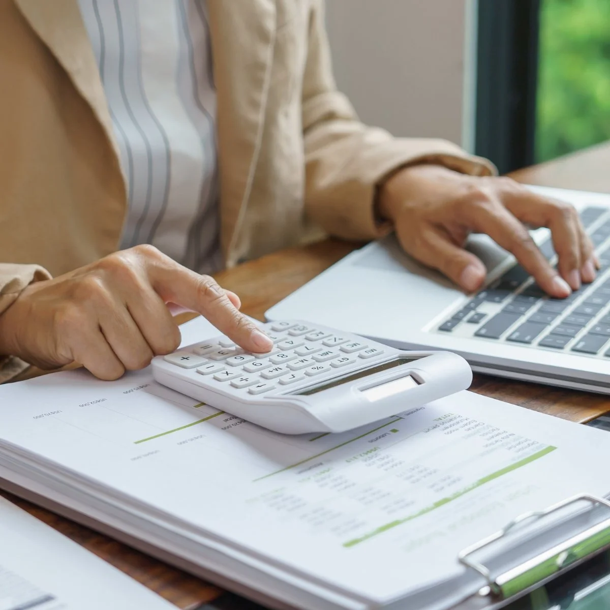 Person using a calculator and laptop on a desk with papers and documents.