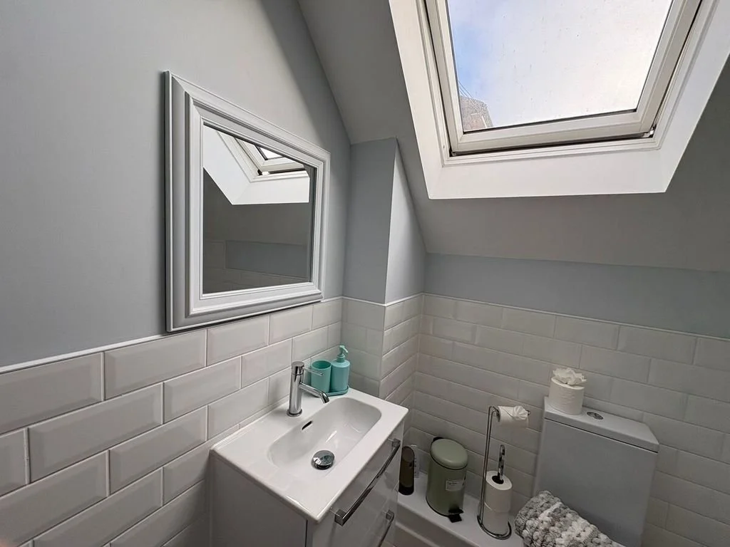 Bathroom with white subway tile wainscoting, a small white sink, a mirror, a sloped ceiling, a skylight window, and a toilet with a roll of toilet paper, a small trash bin, and a towel.