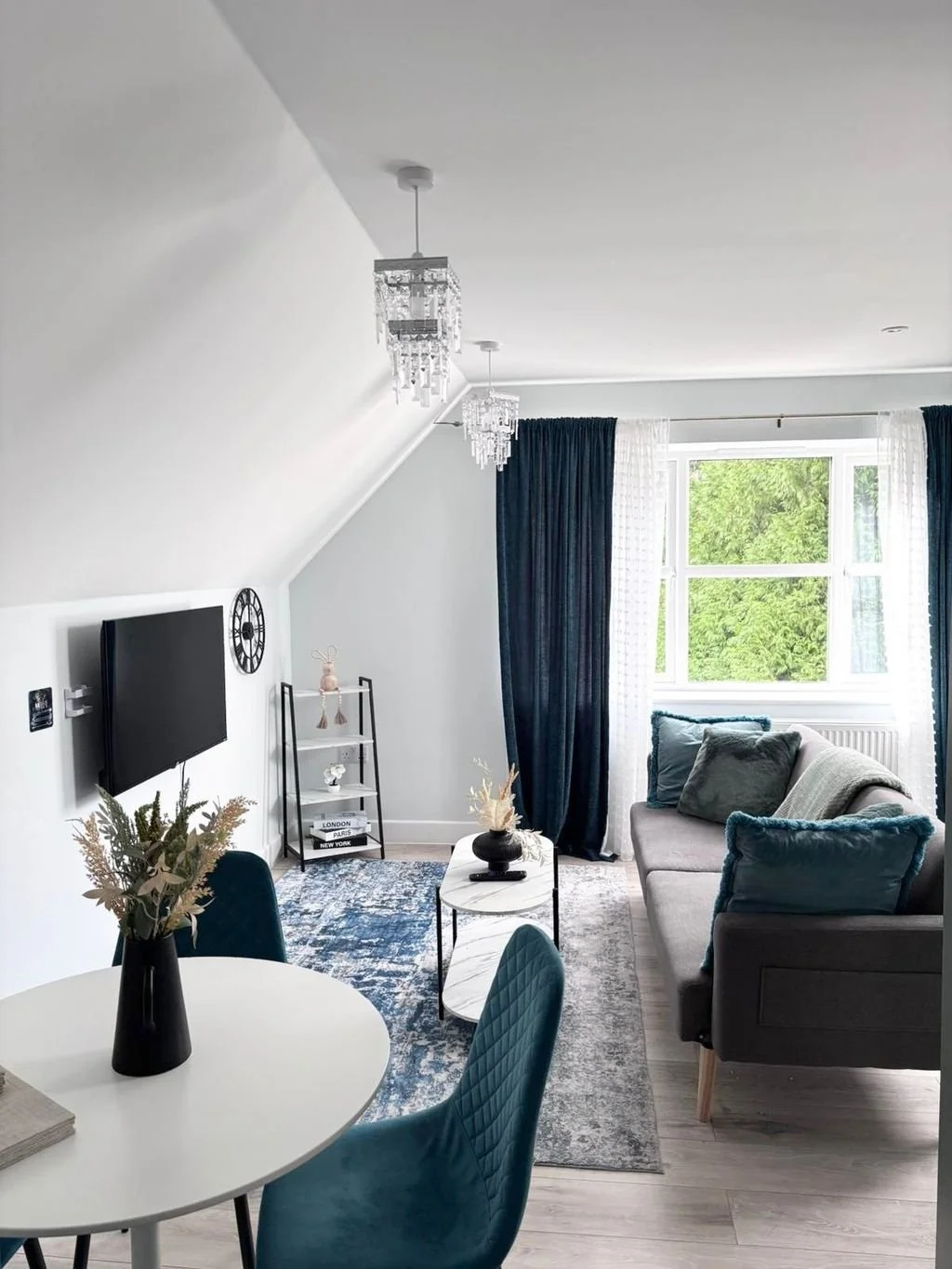 Living room with a gray sofa, navy blue curtains, a window with green trees outside, a white TV mounted on wall, a small white round table with a black vase and plants, a black and white framed shelf, and decorative hanging chandeliers.