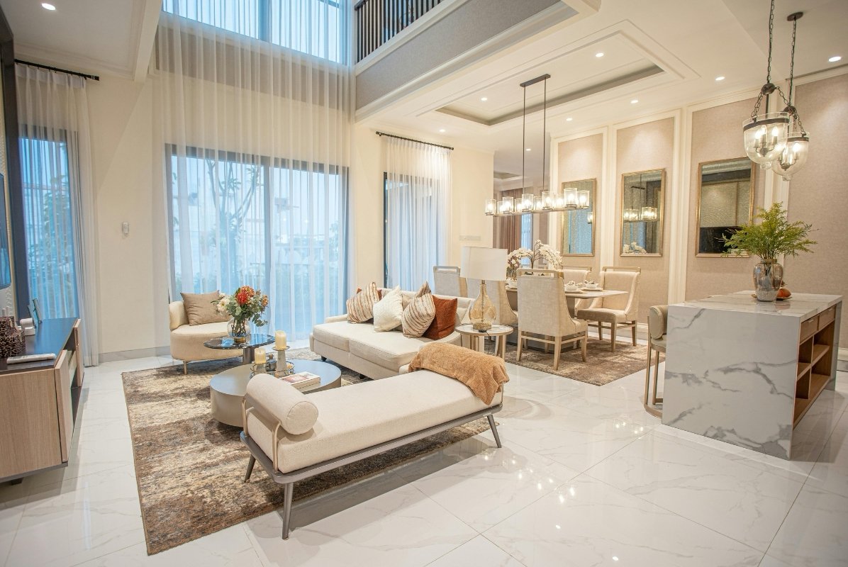 Modern living room and dining area with beige couches, white and glass coffee tables, large windows with sheer curtains, and elegant lighting fixtures.