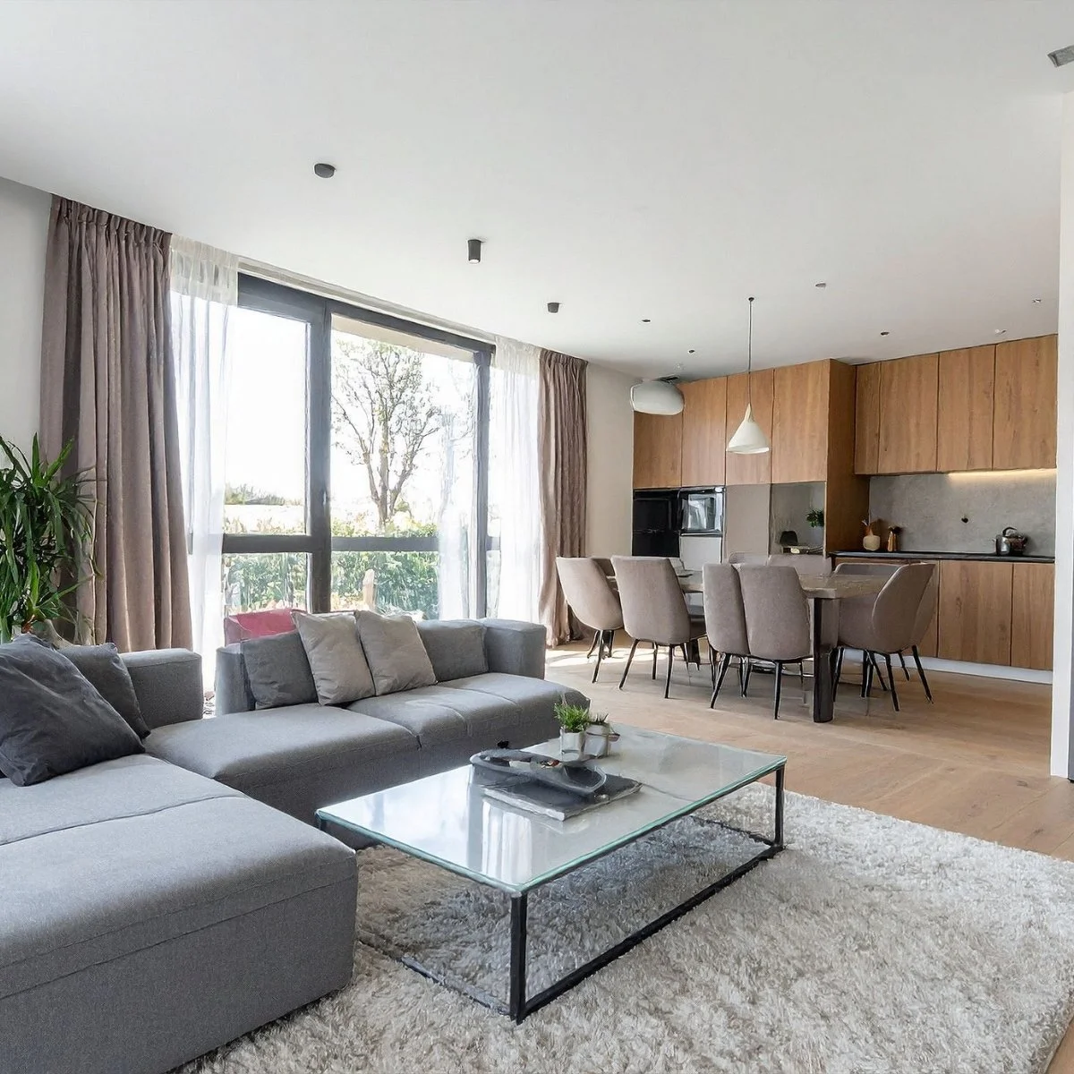 Modern living room and dining area with large windows, gray sofa, glass coffee table, dining table with six beige chairs, wooden kitchen cabinets, and hanging pendant lights.