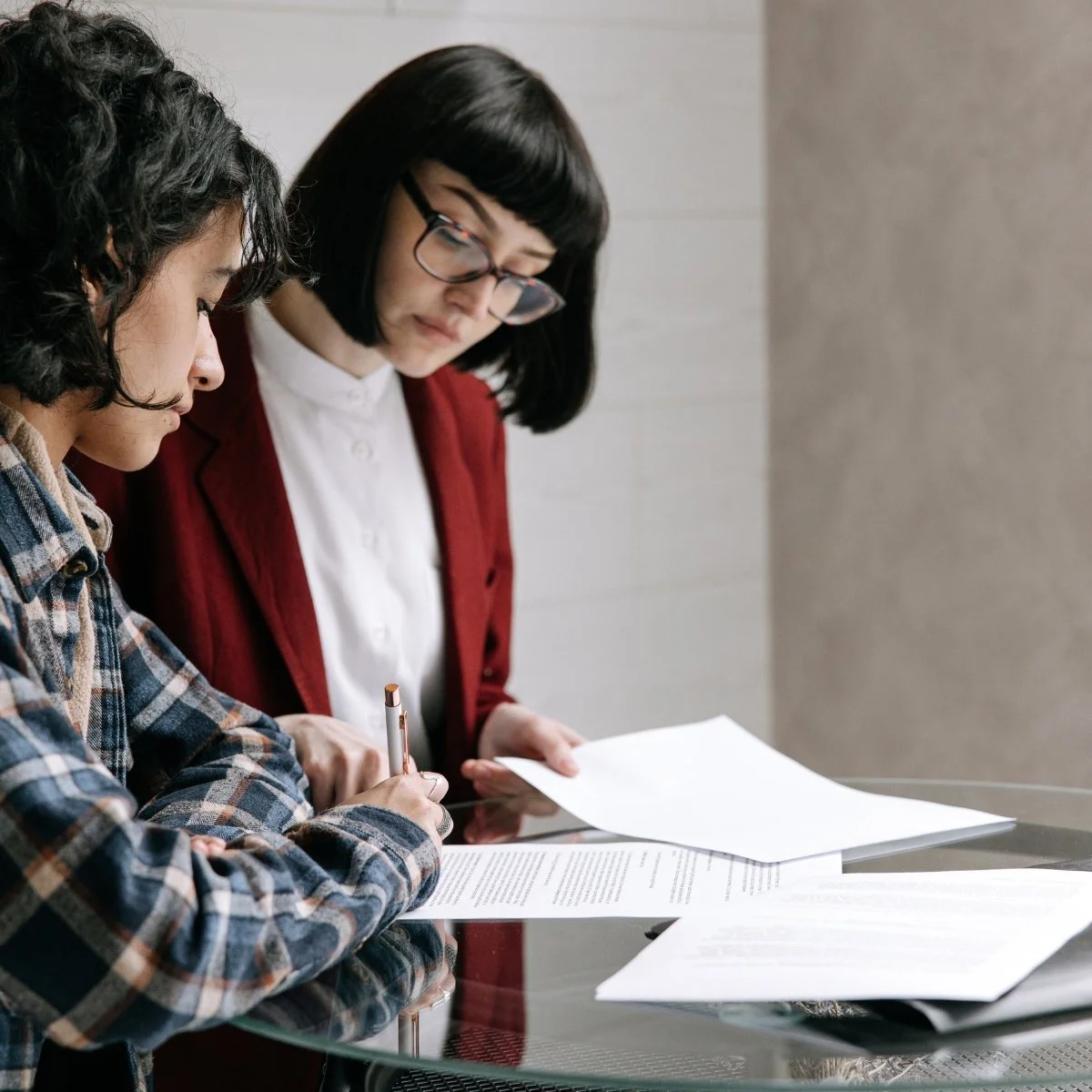 Two women working together at a glass table with papers, one holding a pen, in an indoor setting.