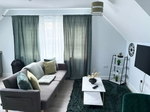Cozy living room with a gray sofa adorned with multicolored pillows, dark green curtains over a large window, a small white coffee table with plants and a remote, a black rug, and a flat-screen TV mounted on the wall.