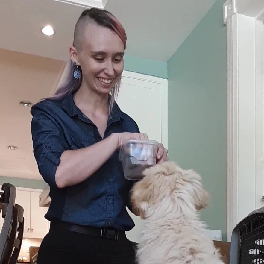 Liz Fisher the dog trainer is wearing a blue shirt, black pants, and blue dangling earrings. They are holding a tupperware of treats and smiling down at a light tan puppy faced away from the camera. They have an undercut and brown and pink hair.