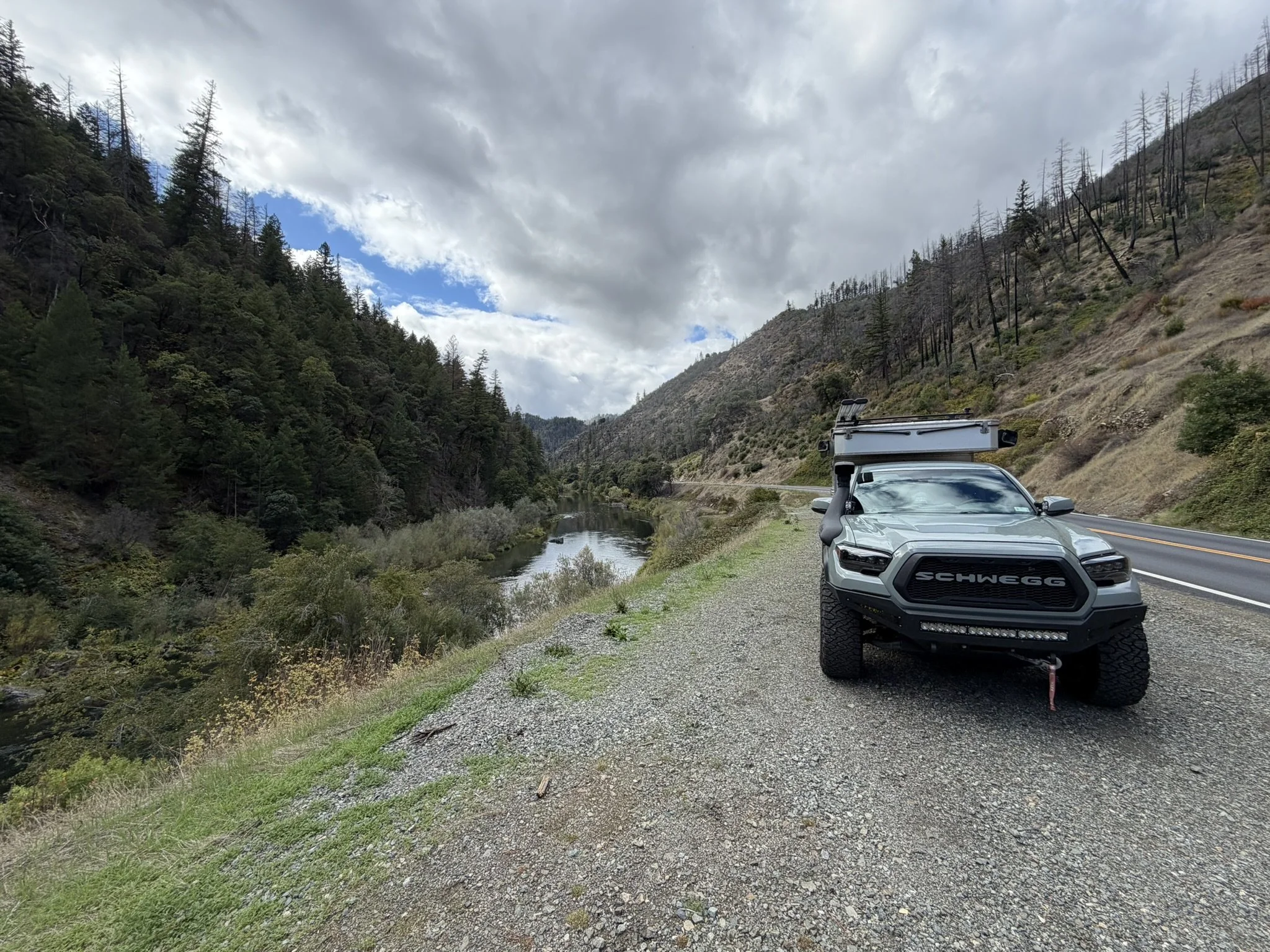 A rugged vehicle is parked beside a river in a canyon, with a mountainous landscape and partially cloudy sky in the background.
