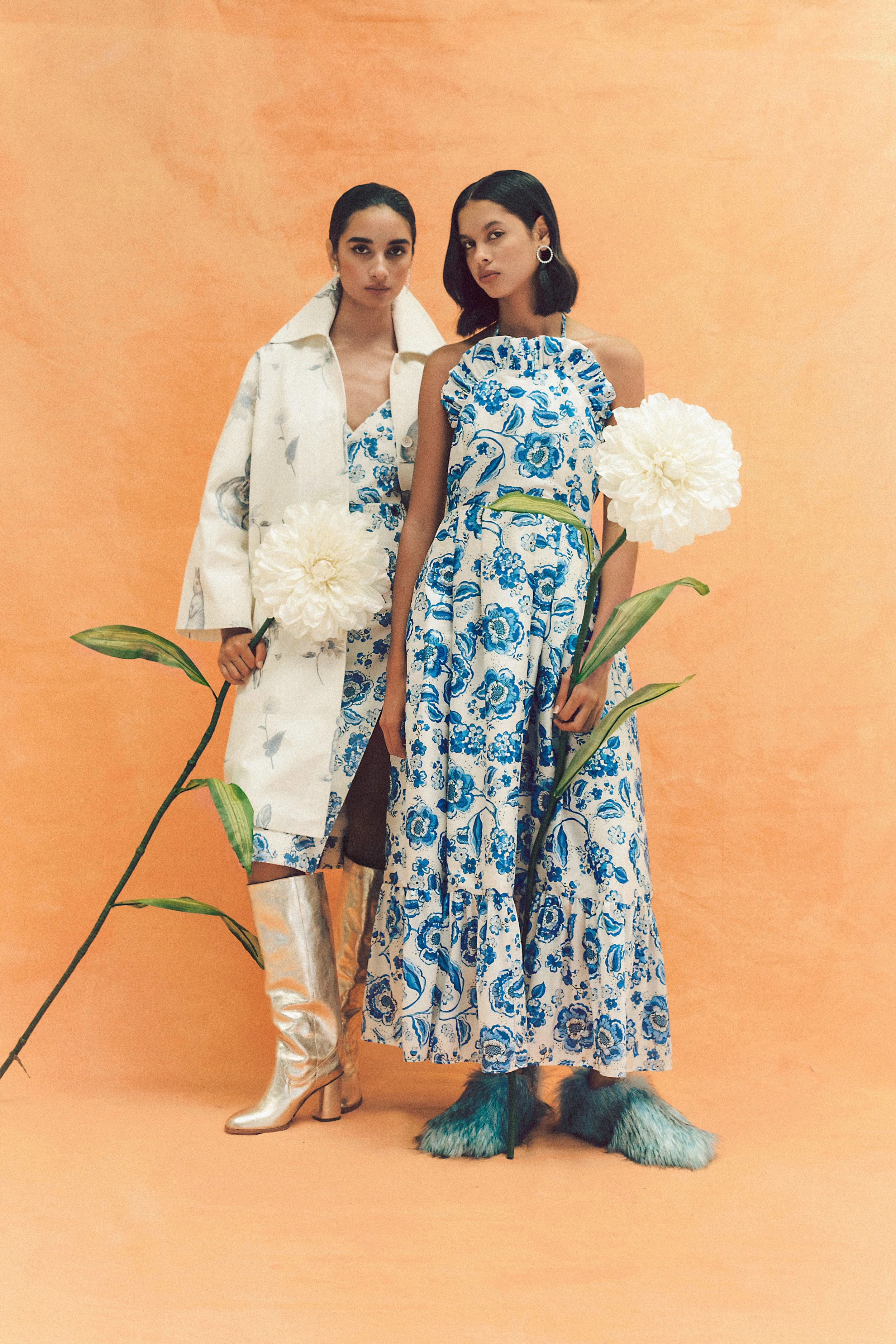 Dois modelos en un fondo de color durazno, una lleva un vestido azul y blanco con florales, y la otra un conjunto blanco con estampados y botas plateadas, sosteniendo flores blancas, acompañadas de un animal con pelaje de colores mezclados en tonos azules, verdes y negros.