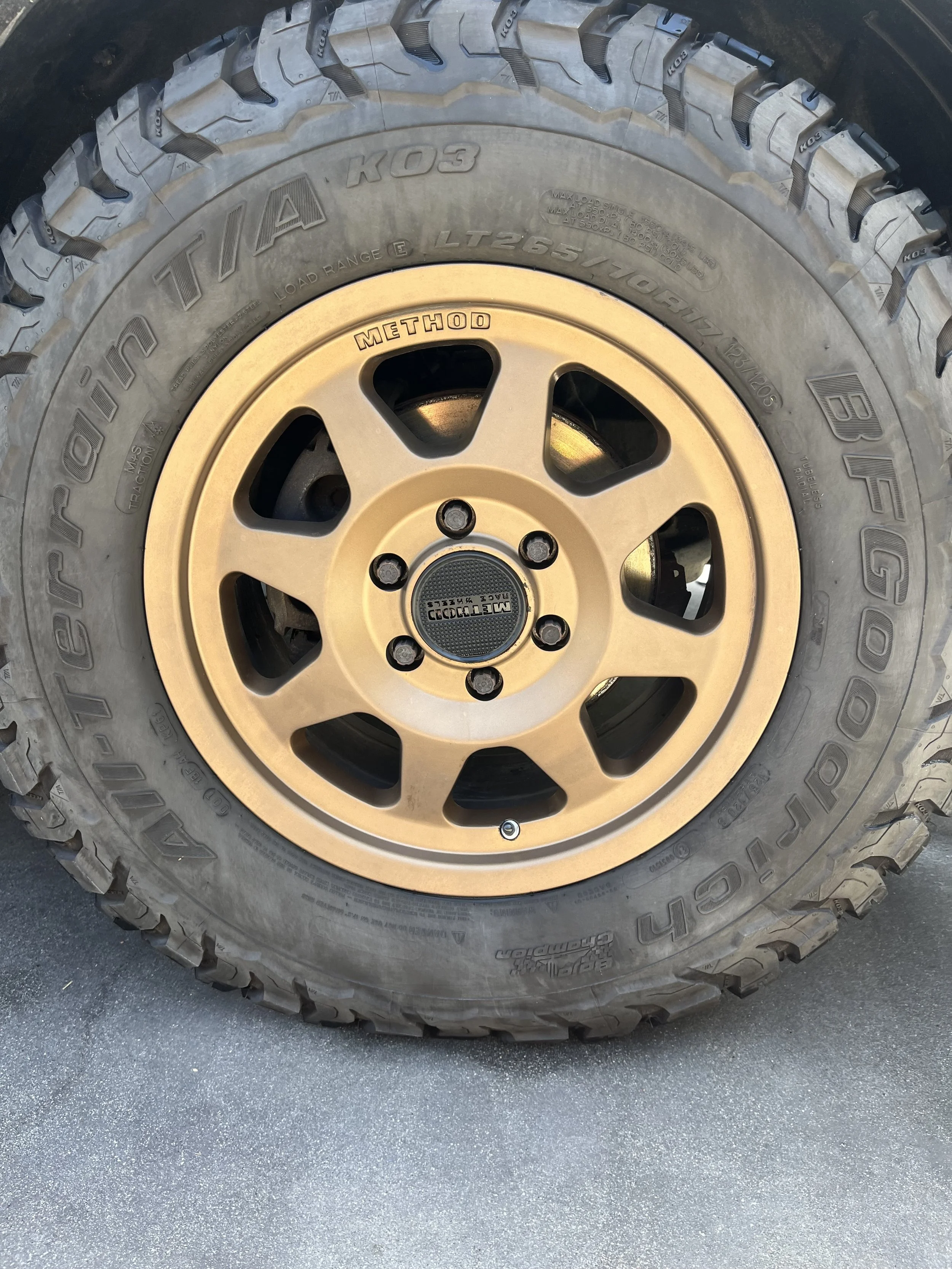 Close-up image of an off-road tire with a tan rim, marked 'BFG KO2' and 'All-Terrain T/A'. The rim has the word 'METHOD' inscribed on one of the spokes. After picture of a van tire during a detail in Del Mar, California.