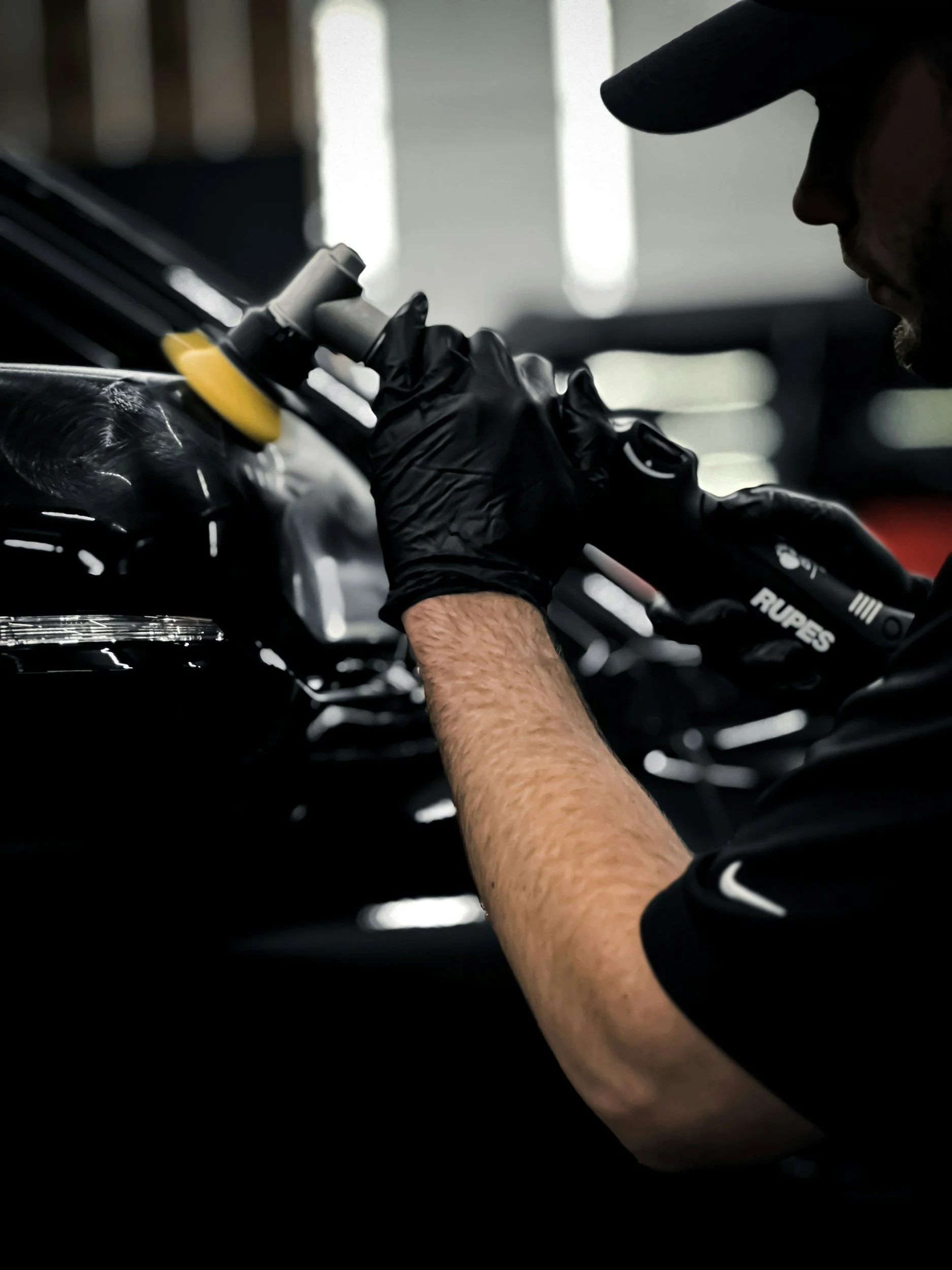 A person using a polishing tool on a black car's surface inside a garage.
