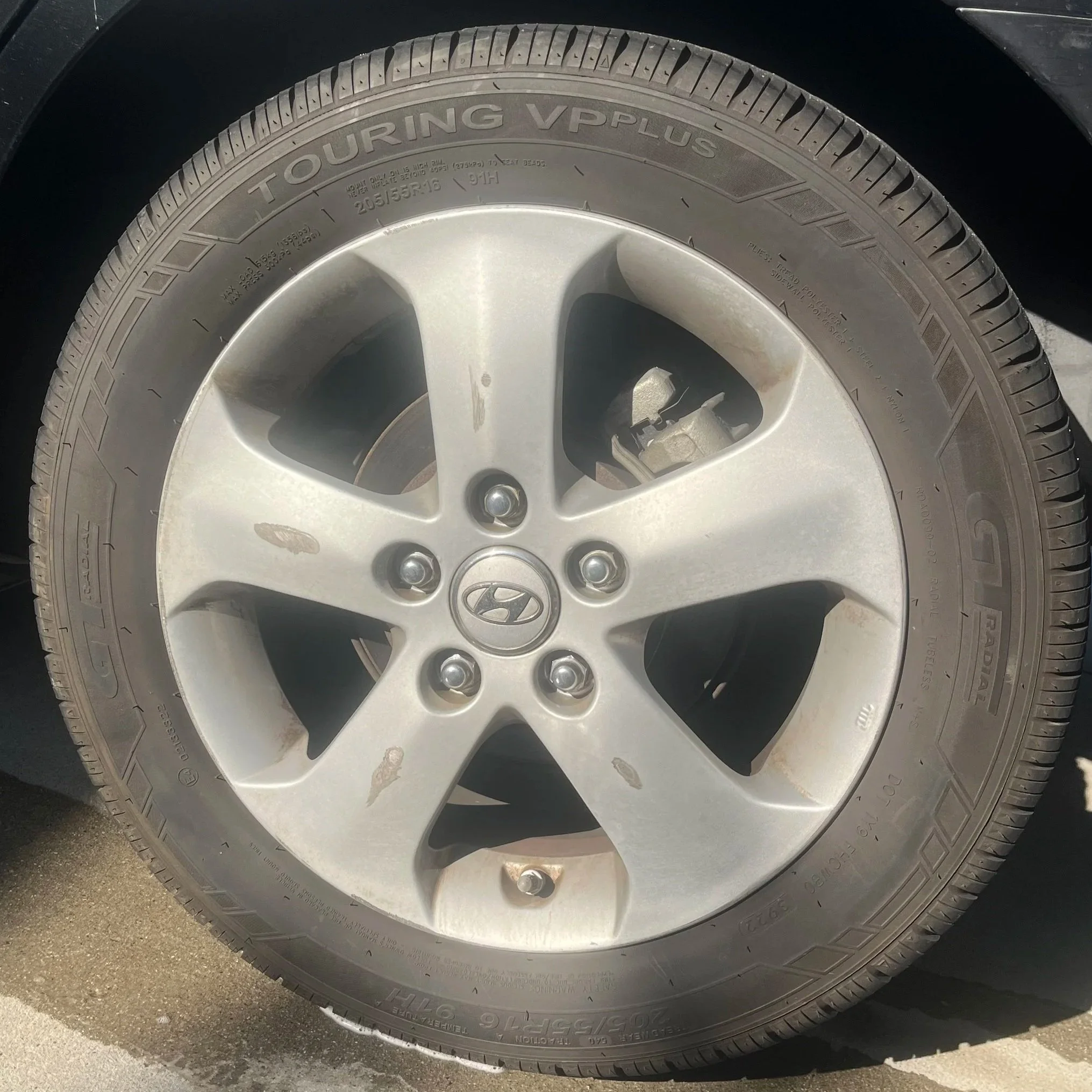 Close-up of a Hyundai car wheel with a silver rim and black Bridgestone tire, showing some dirt and light scratches. Before picture of a car tire during a detail in Torrey Del Mar, California.
