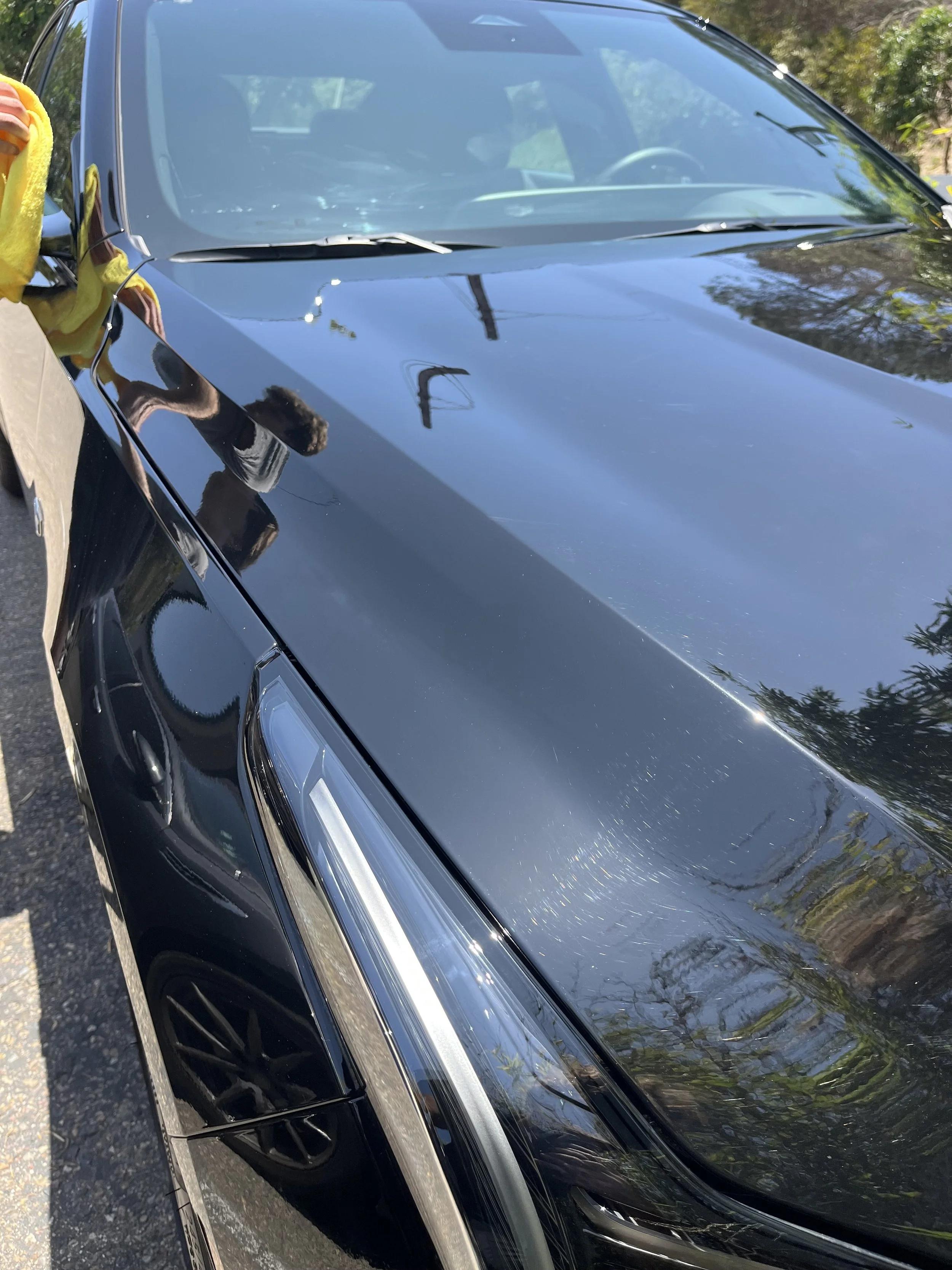 Close-up of a black car's front and side, reflecting trees, power lines, and the sky, parked next to a person wearing a yellow jacket. An after photo of an exterior detail in San Deigo.