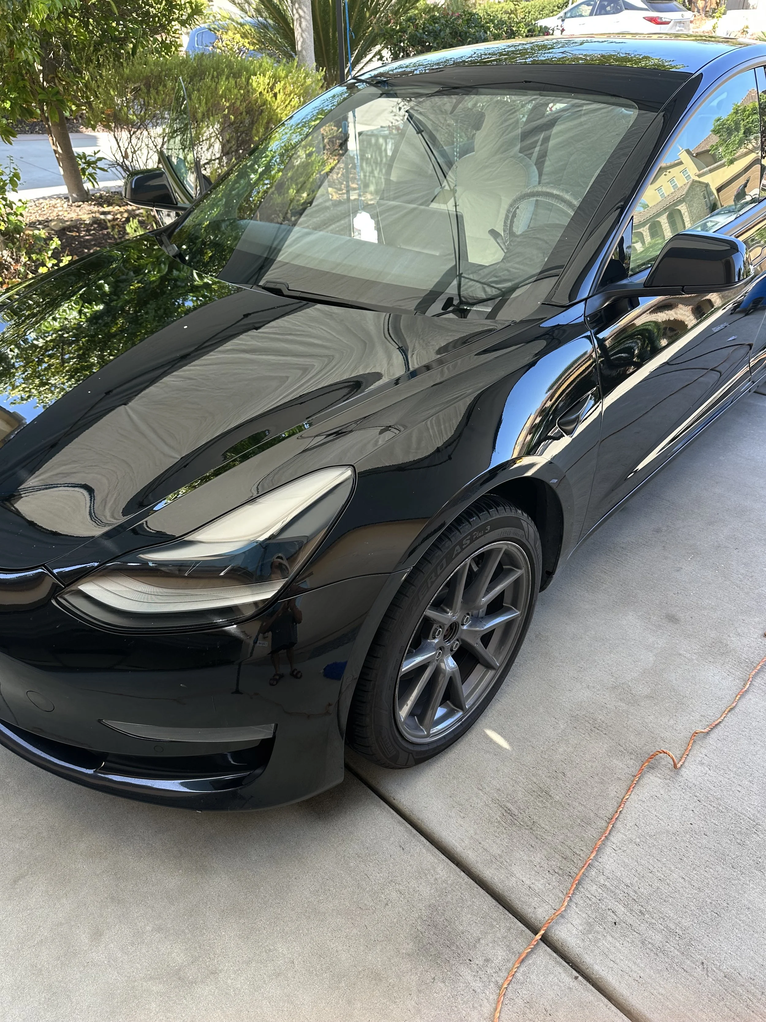 A black sports car parked on a concrete driveway, reflected in the shiny surface of the car. The car has modern headlights, dark gray wheels, and side mirrors. Surrounding greenery and trees are visible in the background, along with other parked cars and a house.