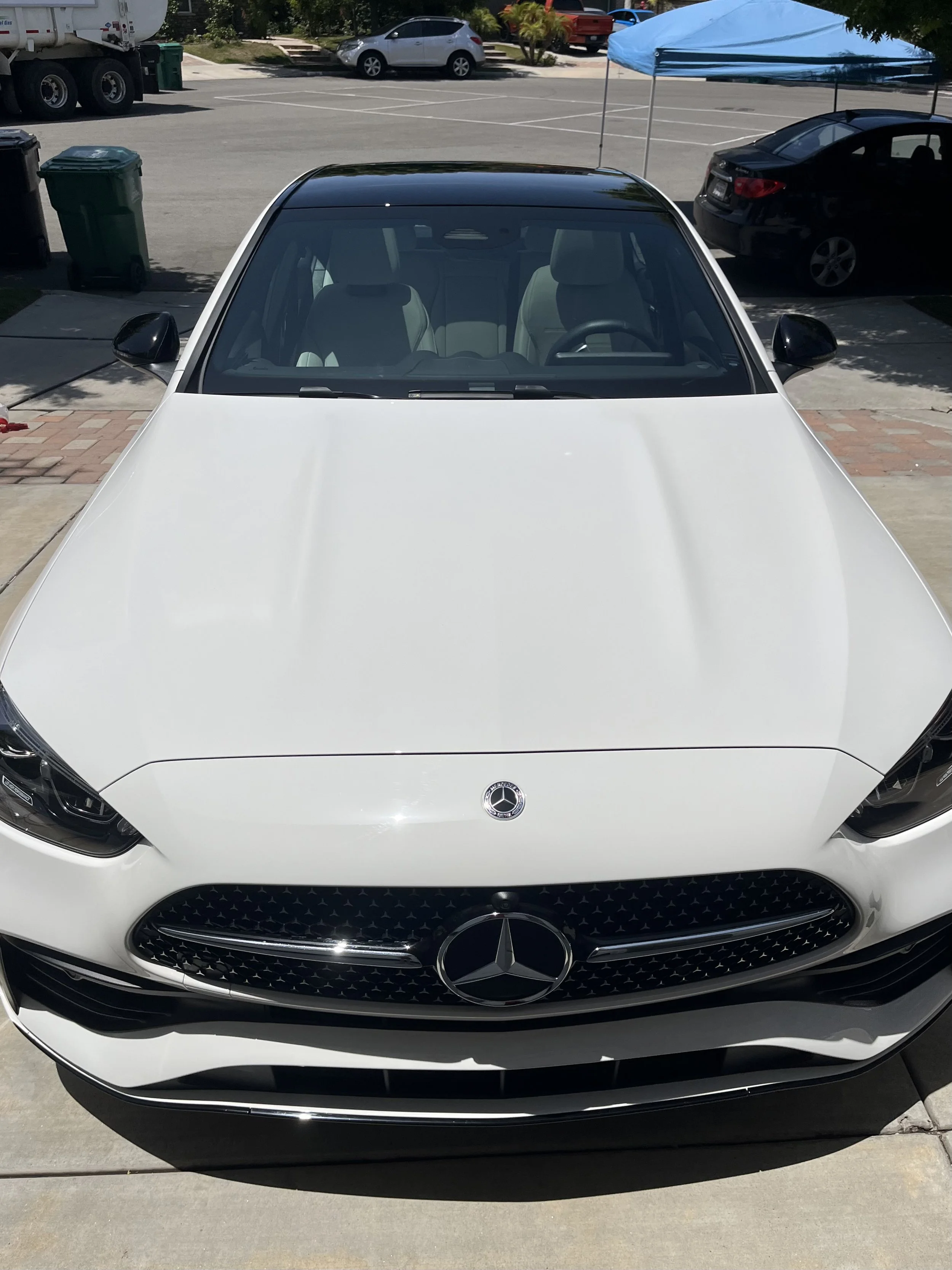Front view of a white Mercedes-Benz car parked in a driveway, with a parking lot and other cars in the background. An after photo of an exterior detail in Torrey Pines, San Diego