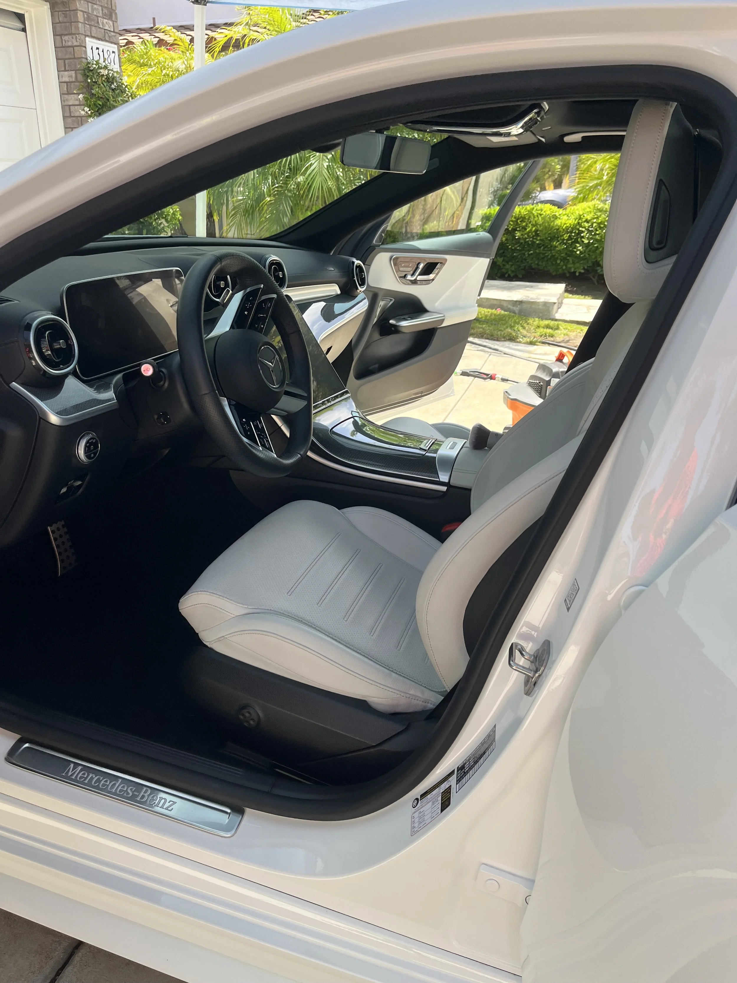 The interior of a white Mercedes-Benz car showing the driver's seat, steering wheel, dashboard, and part of the front passenger seat with a light-colored headrest, set against a background of greenery and a driveway.
